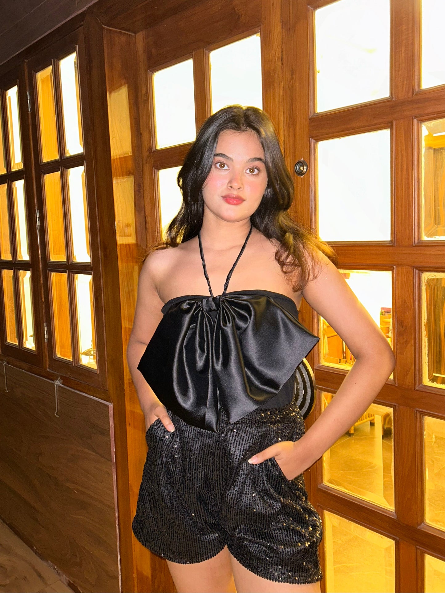 Model wearing Luna high waisted shorts in black sequins with satin bow halter top by wooden window