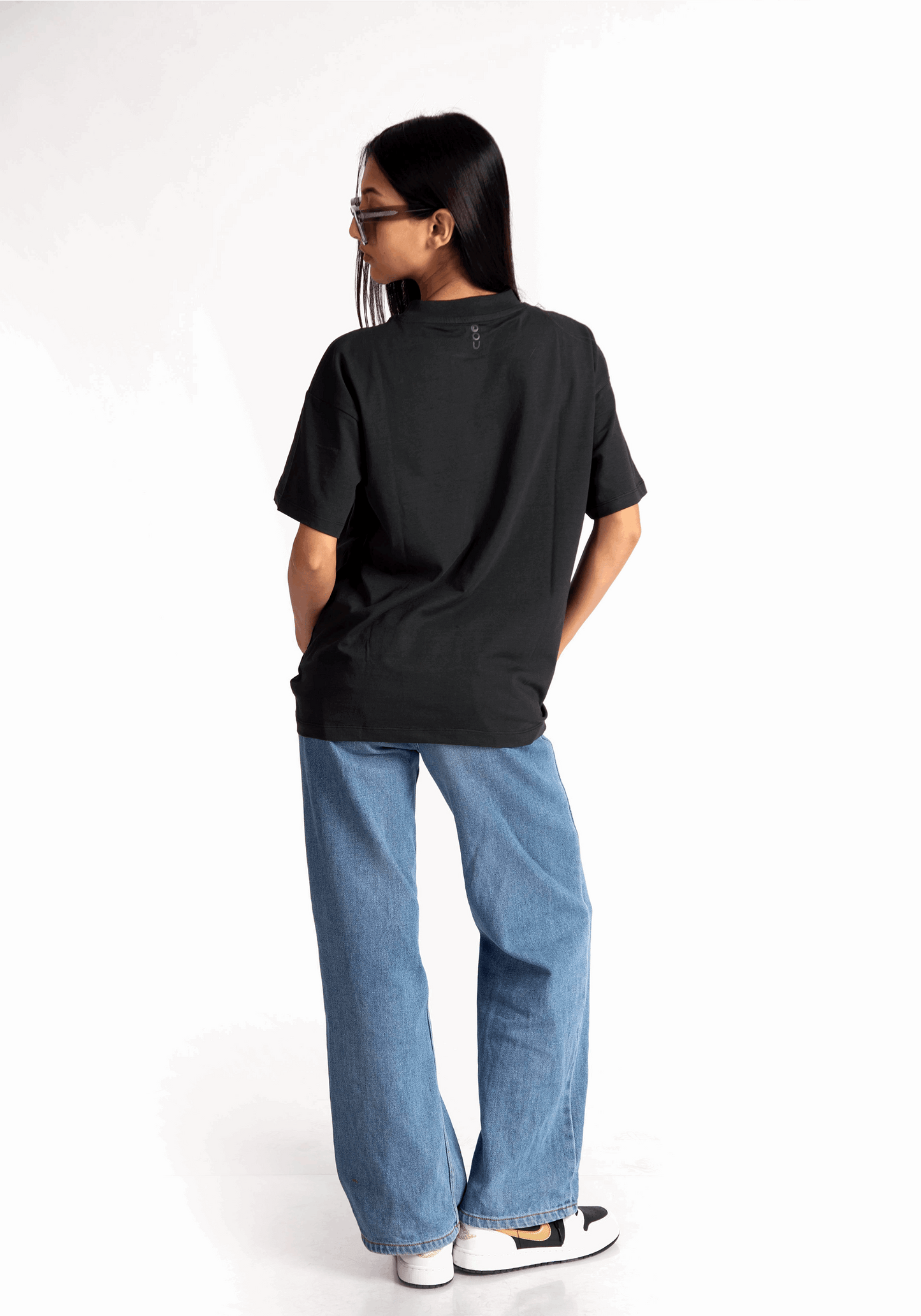 Back view of a woman wearing Stylera women's relaxed fit black tee paired with blue jeans and white sneakers