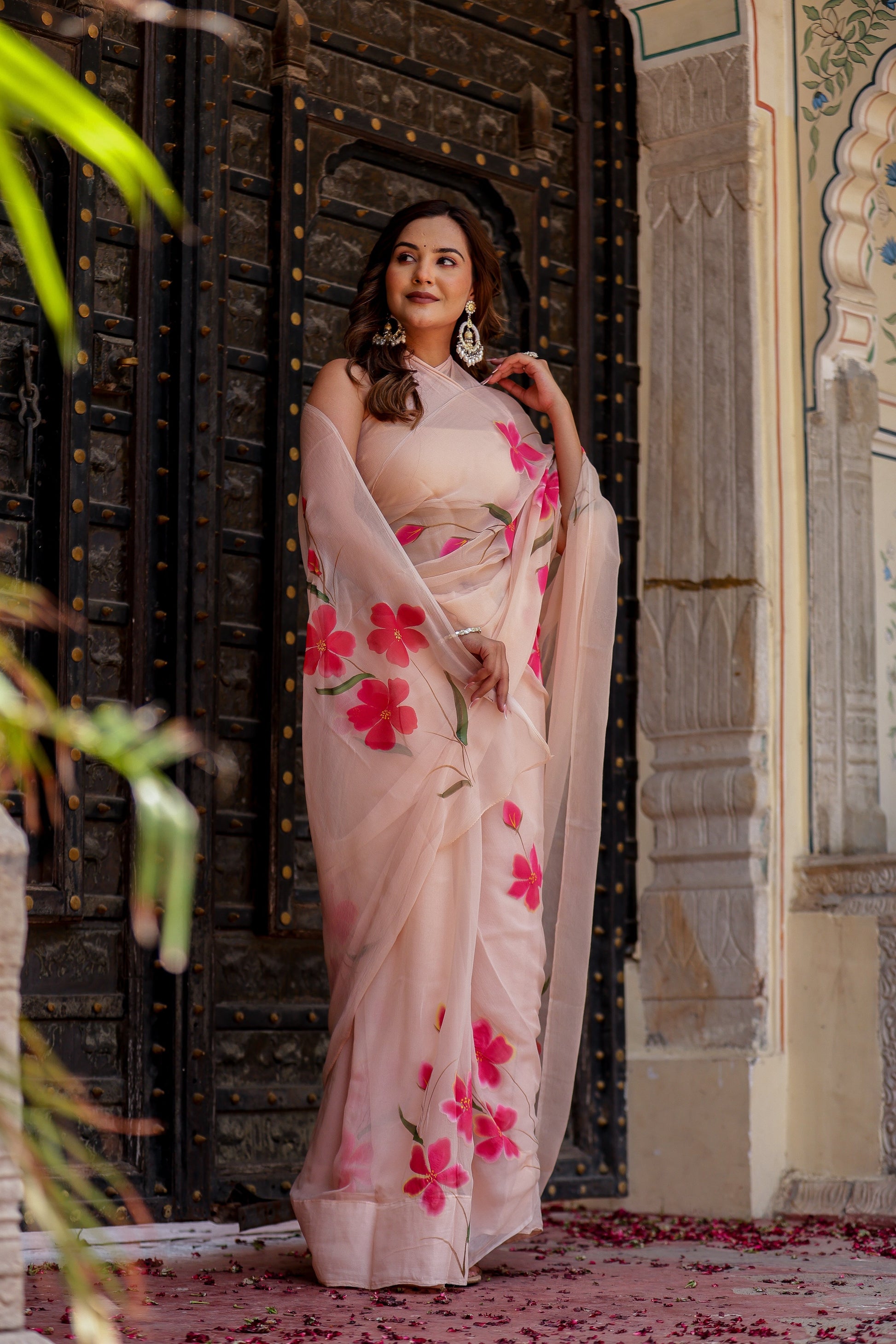 Model wearing a beige floral chiffon saree adorned with vibrant pink flowers, styled elegantly outdoors