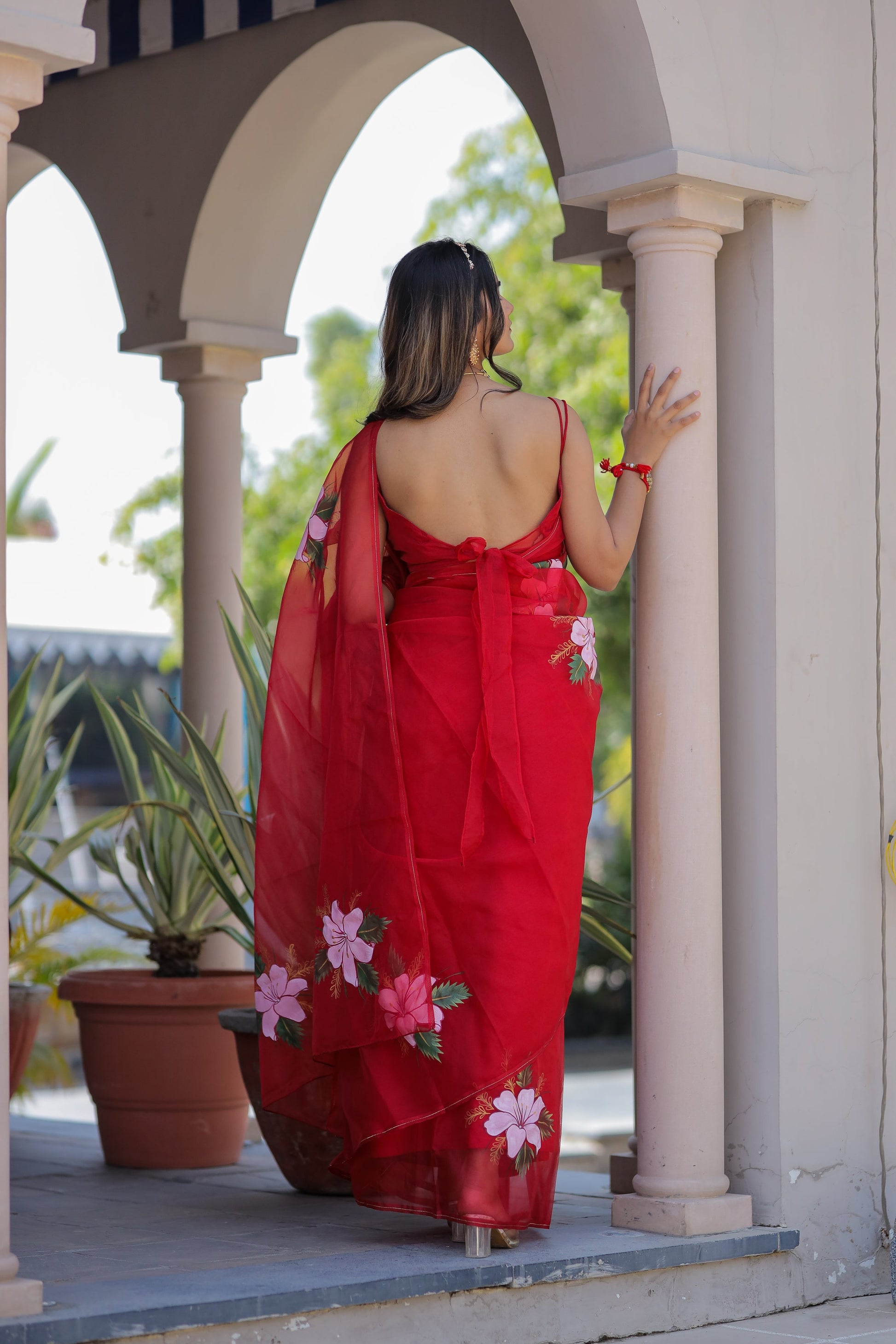 Elegant handpainted red organza saree with floral designs, showcased outdoors for a feminine look