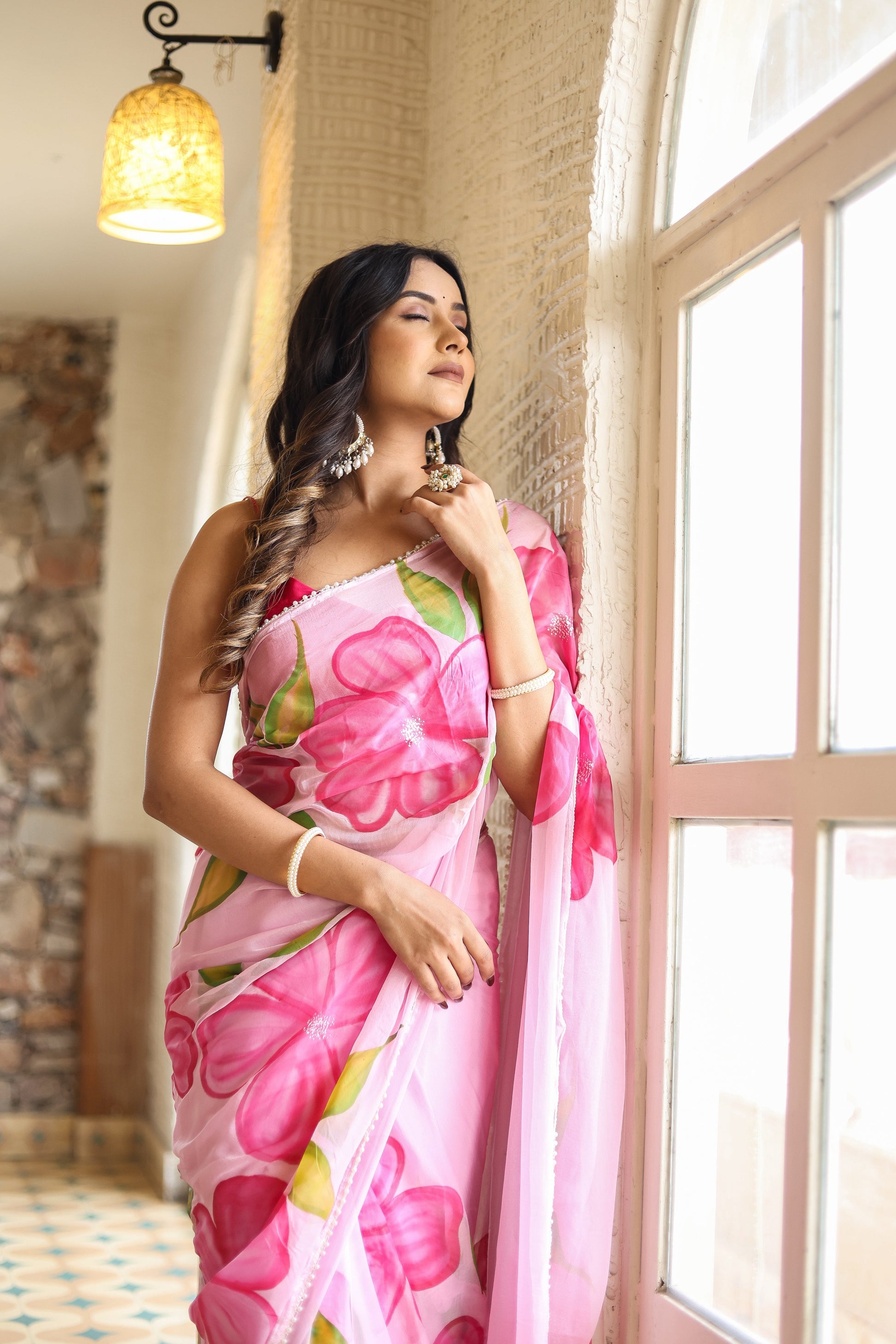 Lightweight party wear saree in pink with floral design, elegantly worn by a model near a window