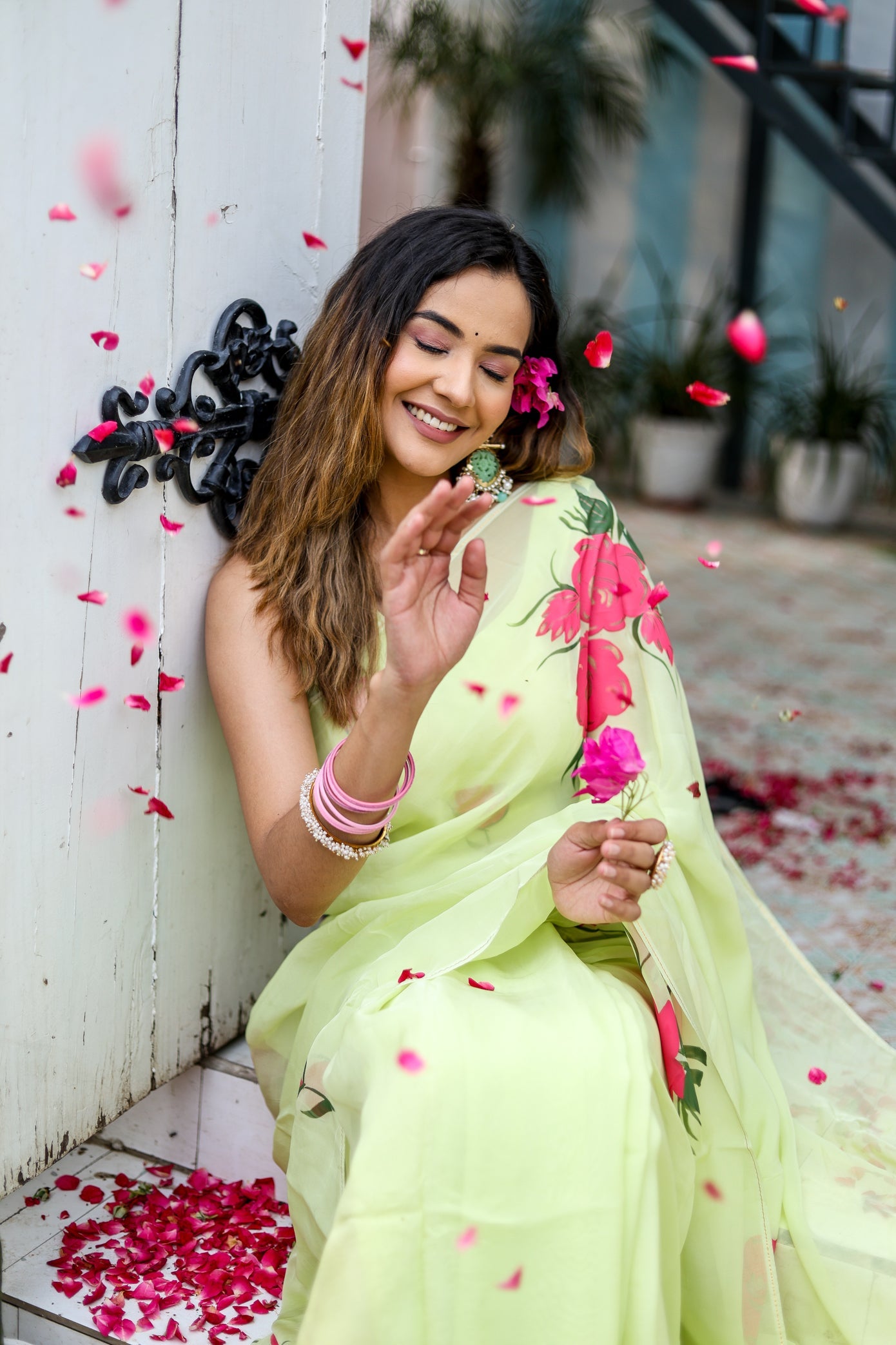 Model wearing a lightweight pastel saree with floral print, surrounded by flower petals in a vibrant setting