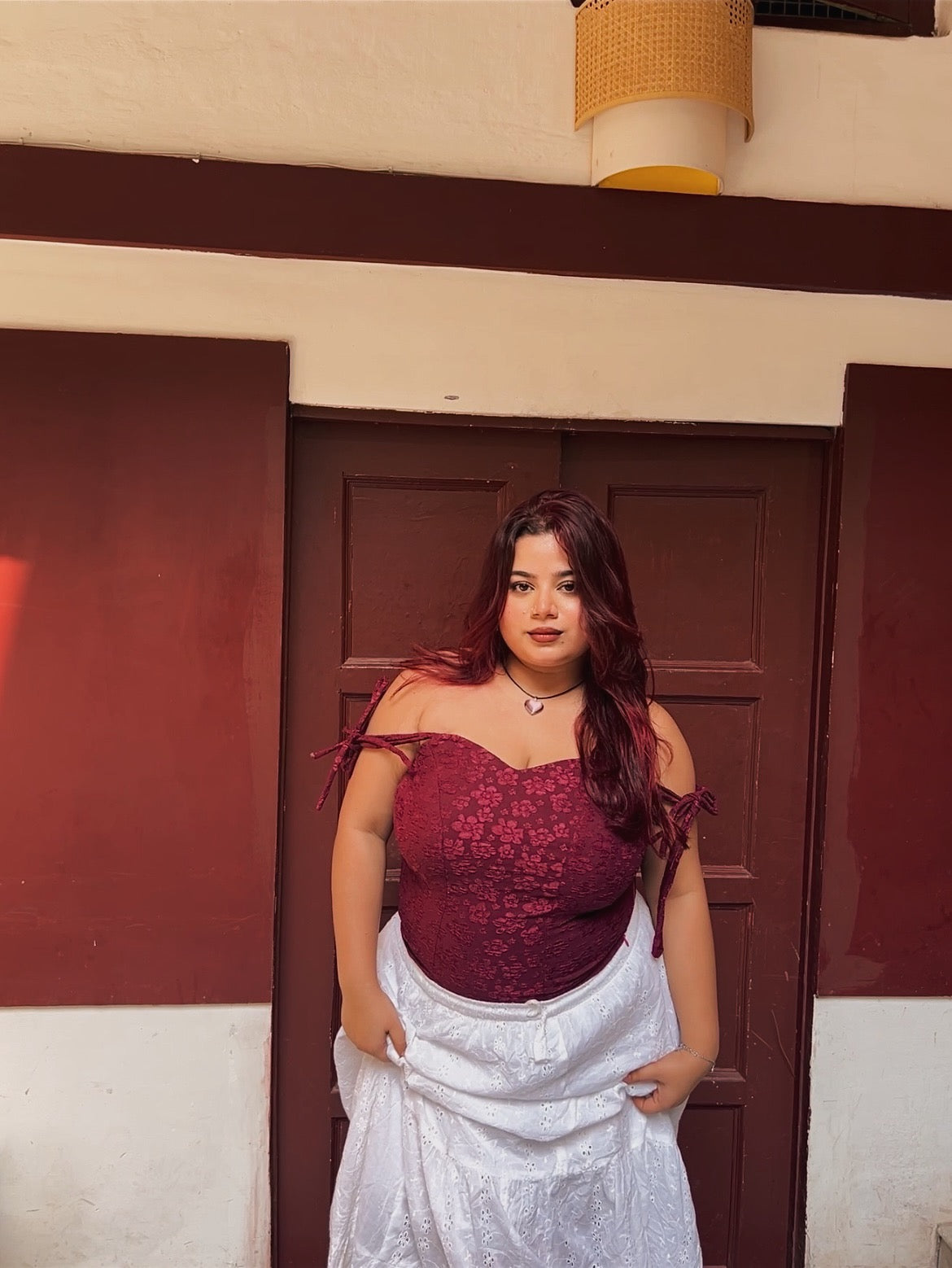 Model wearing red lace corset top for women with white skirt standing against a brown door backdrop