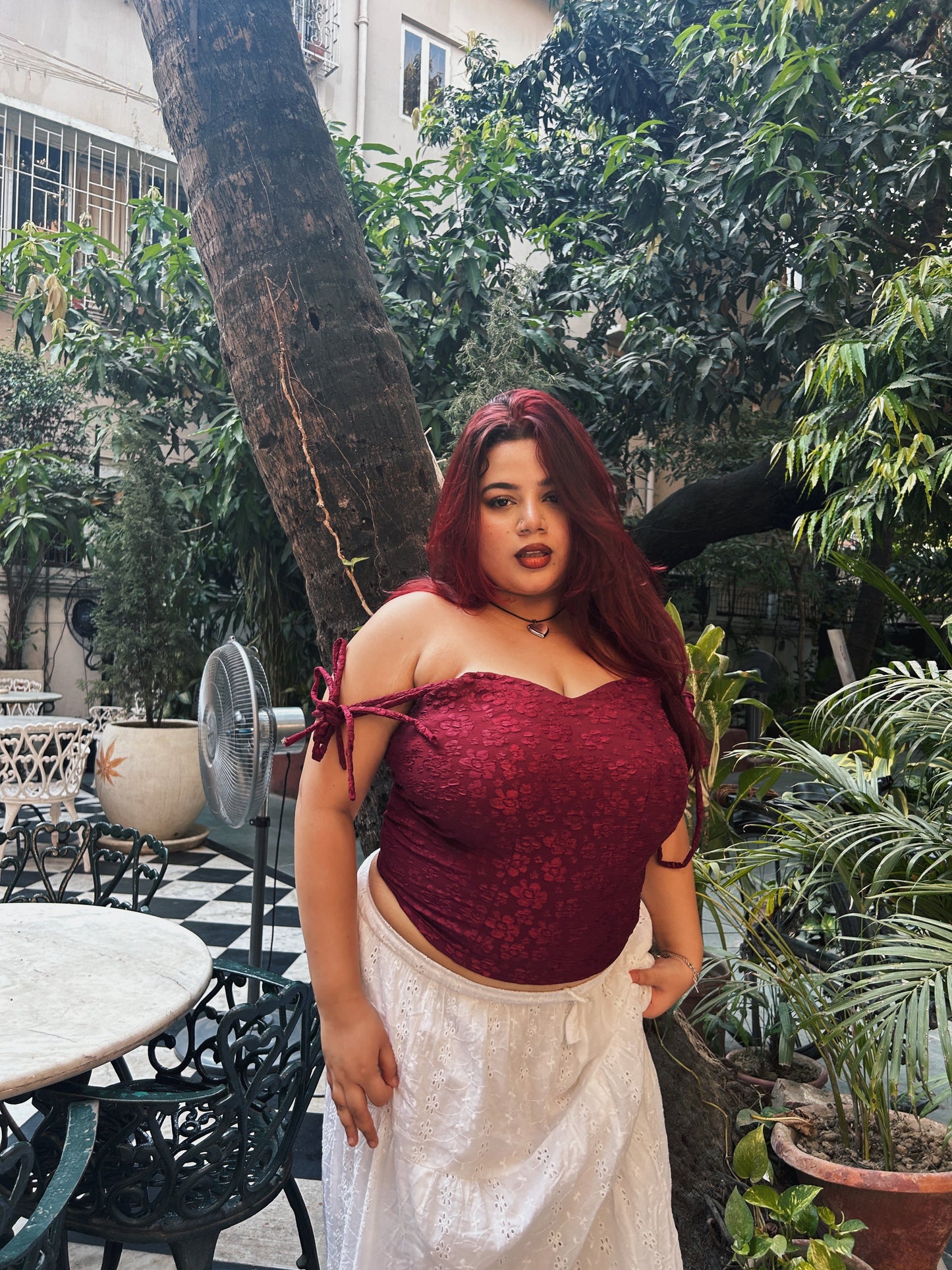 Plus size model wearing a red lace corset top for women with off shoulder tie straps and white skirt in a garden patio