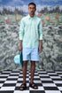 Male model wearing malmal regular fit formal shirt in mint green with striped blue shorts and hat on checkered floor