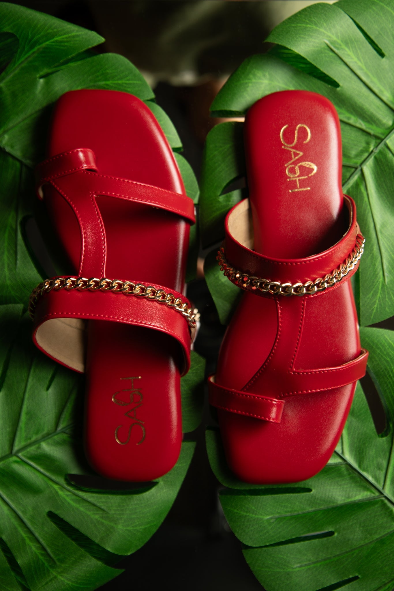 Stylish red flat sandals by Stylera with gold chain detail on green leaves background