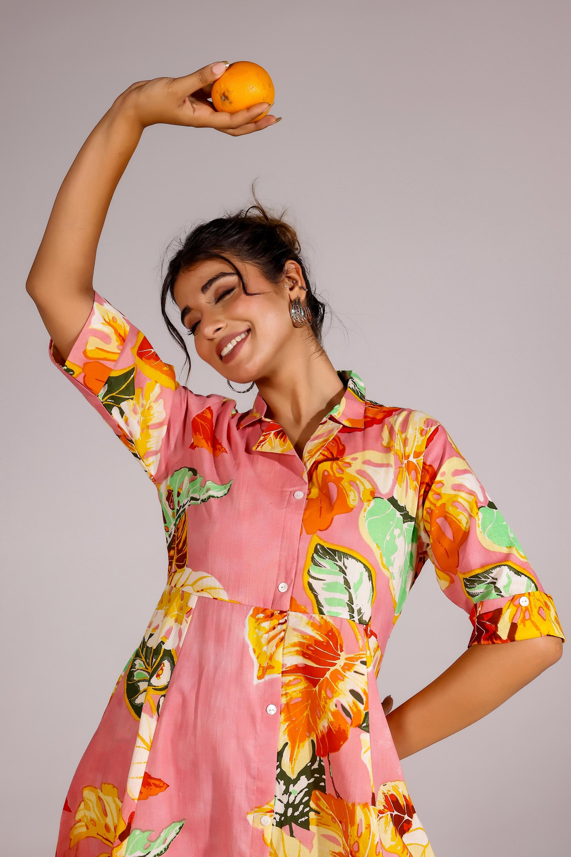 Model wearing a summer pink cotton dress with vibrant floral patterns, holding an orange fruit