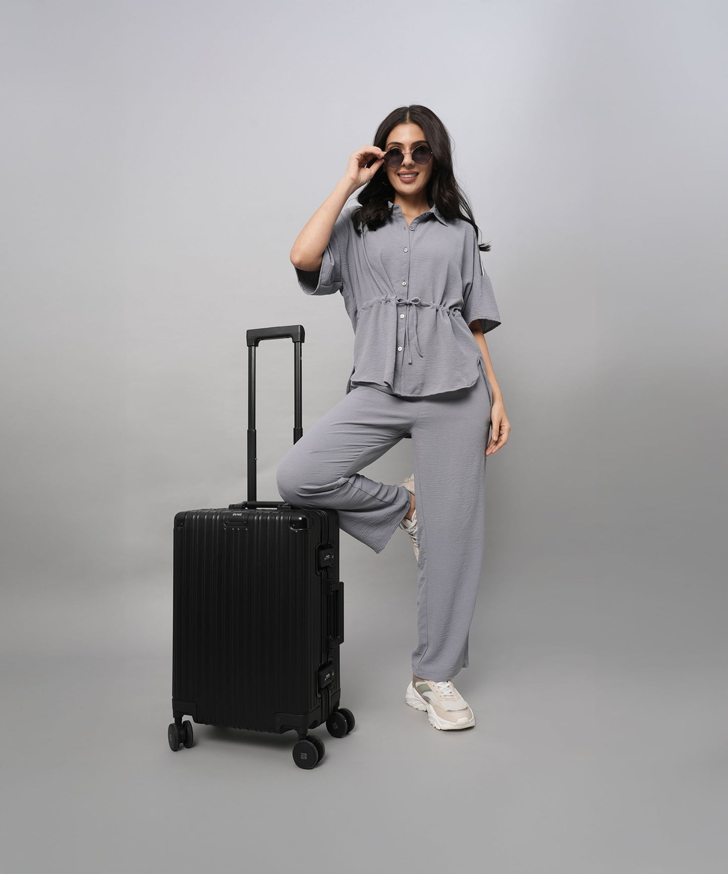 A woman poses confidently in a Grey Tie Up Top with Loose Pants Cord Set and sunglasses, one foot on a black suitcase, against a grey background.