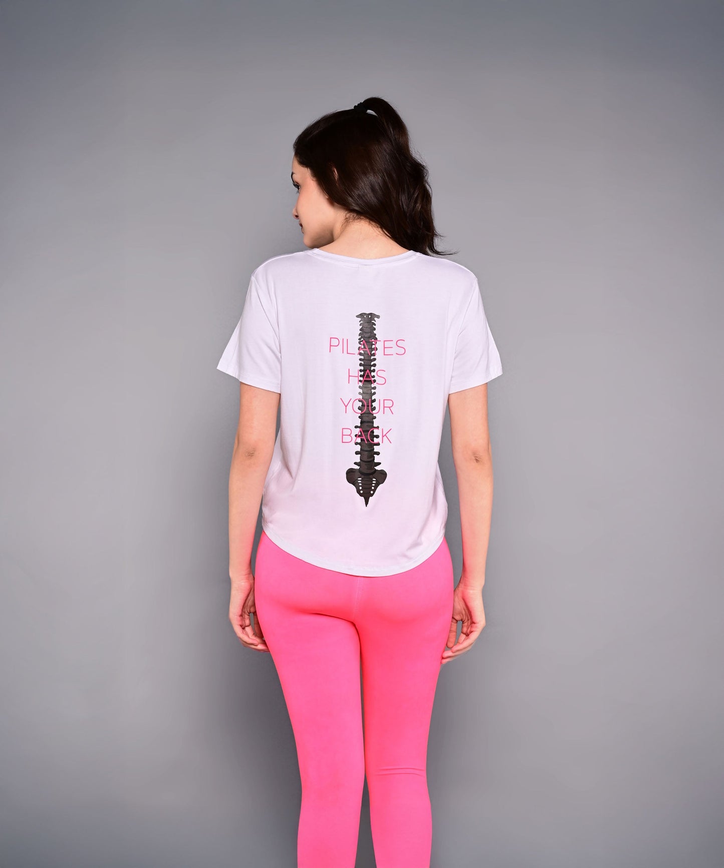 Wearing the 'Pilates Has Your Back' Printed Tee, this woman sports pink leggings and a white cotton-modal shirt with a spine graphic—an ideal motivational top for her next workout.