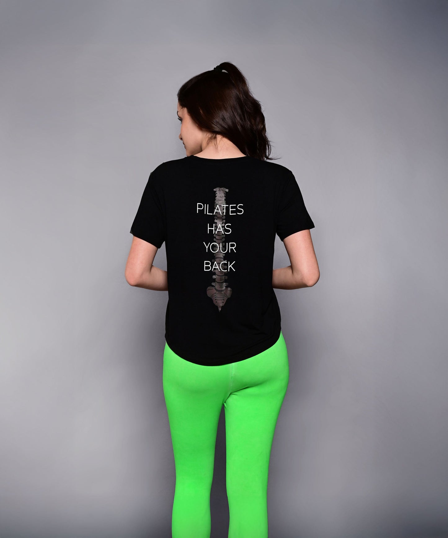 A woman in bright green leggings wears the 'Pilates Has Your Back' Printed Tee, featuring the motivational slogan on the back—perfect for workouts or casual wear.