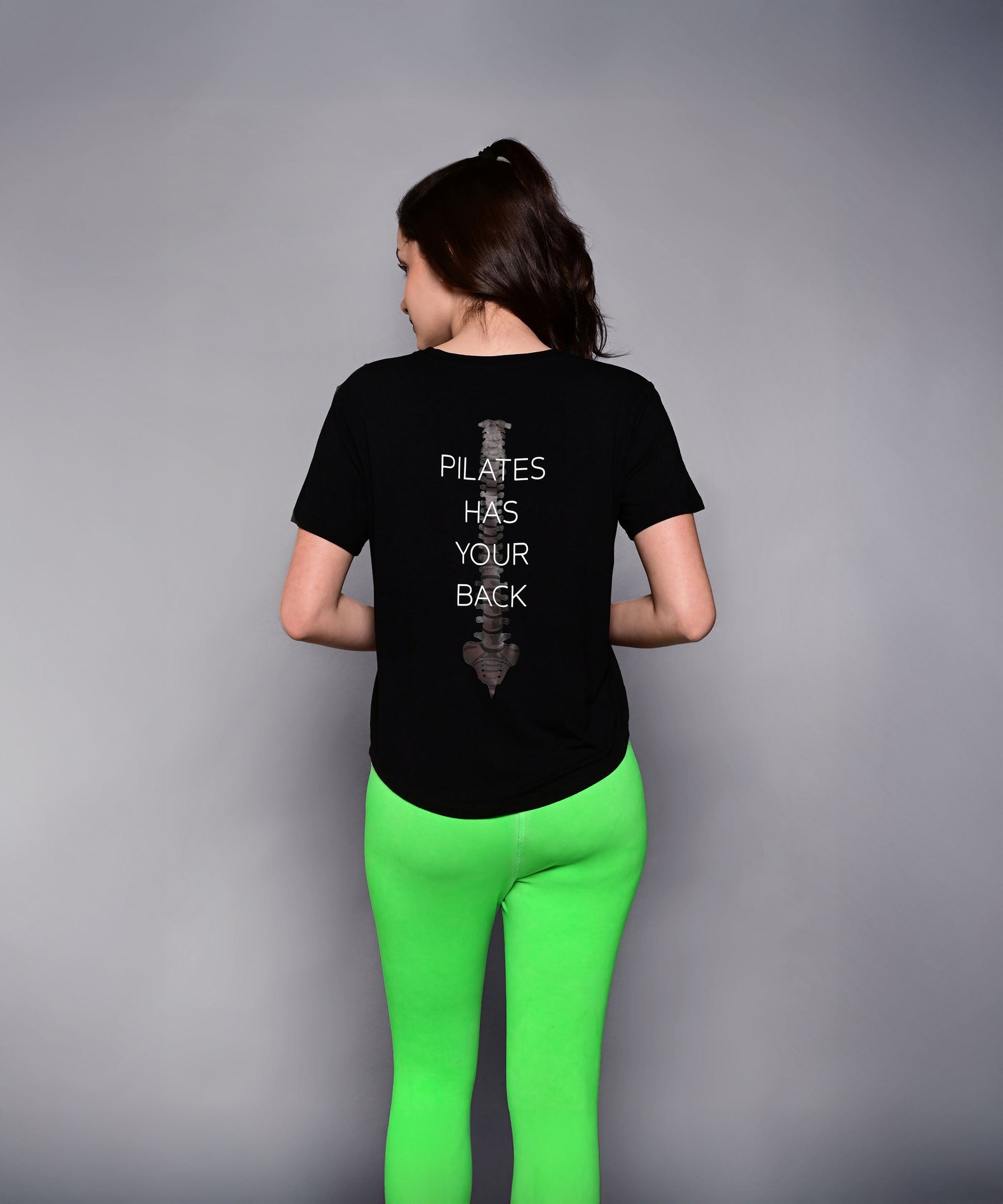 A woman in bright green leggings wears the 'Pilates Has Your Back' Printed Tee, featuring the motivational slogan on the back—perfect for workouts or casual wear.