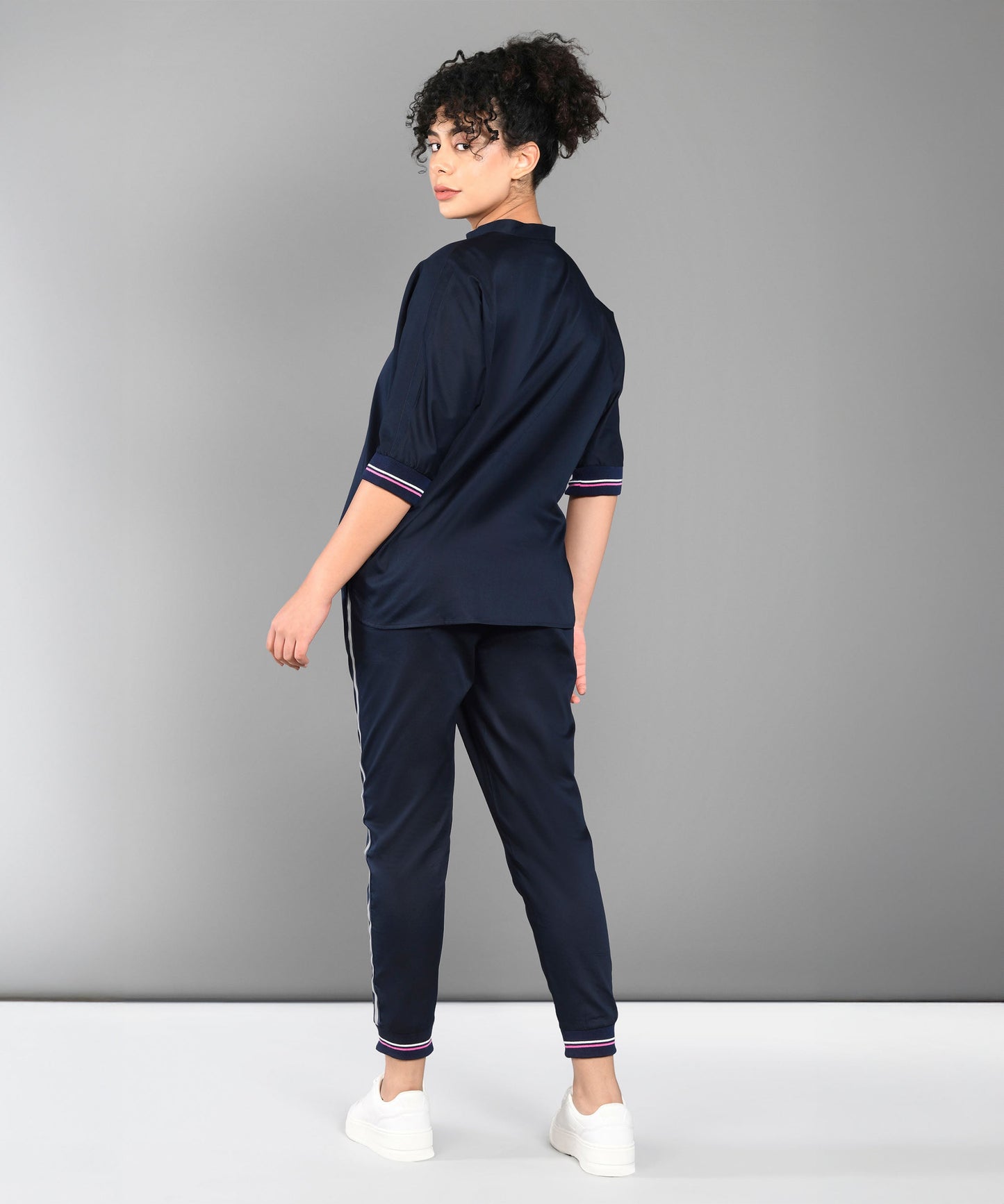 A woman in the Accented Set, a navy blue cotton co-ord, stands facing away and turns her head toward the camera, paired with white sneakers.