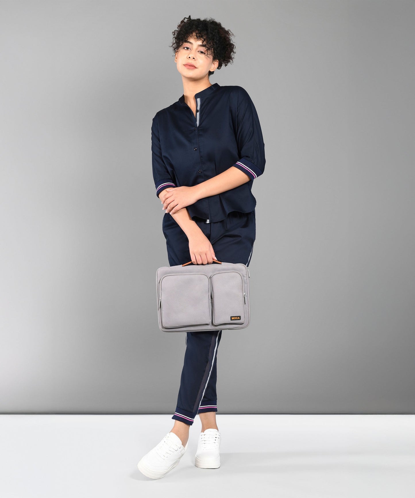 A person wears the Accented Set in navy blue cotton, paired with white shoes and holding a gray bag, standing before a plain gray background.