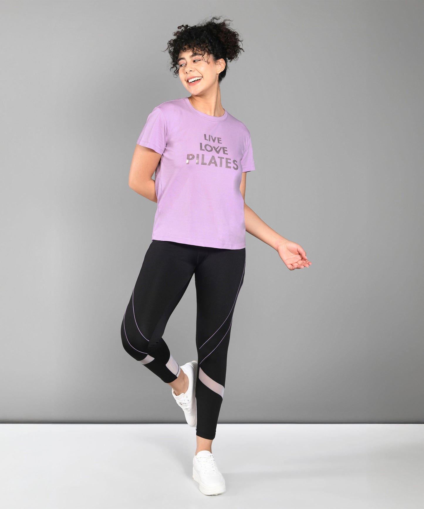 Woman in activewear smiling, wearing a Pilates Pink T-shirt, black leggings, and white sneakers.