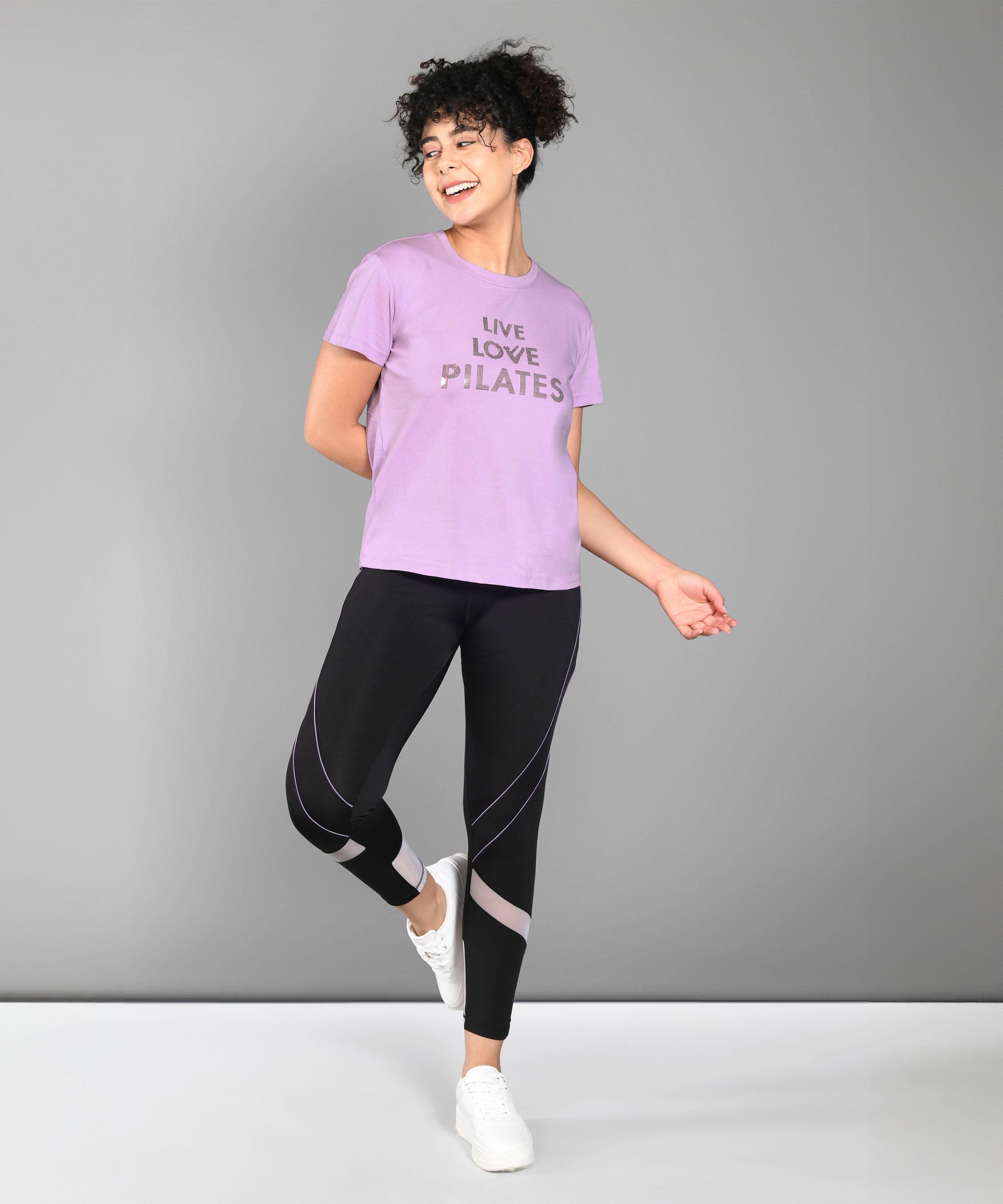Woman in activewear smiling, wearing a Pilates Pink T-shirt, black leggings, and white sneakers.