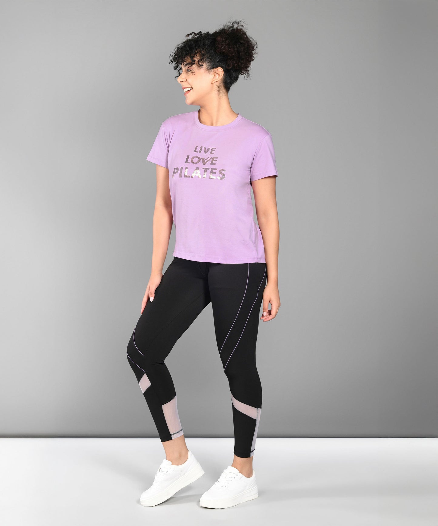 Woman in a Pilates Pink T-shirt, black leggings, and white sneakers standing and smiling.