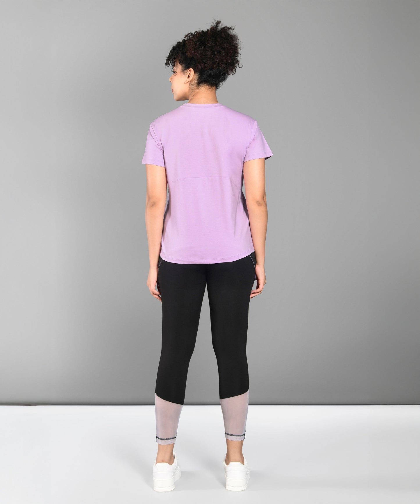 A woman faces away, wearing a Pilates Pink T-shirt, black leggings with gray cuffs, and white sneakers.