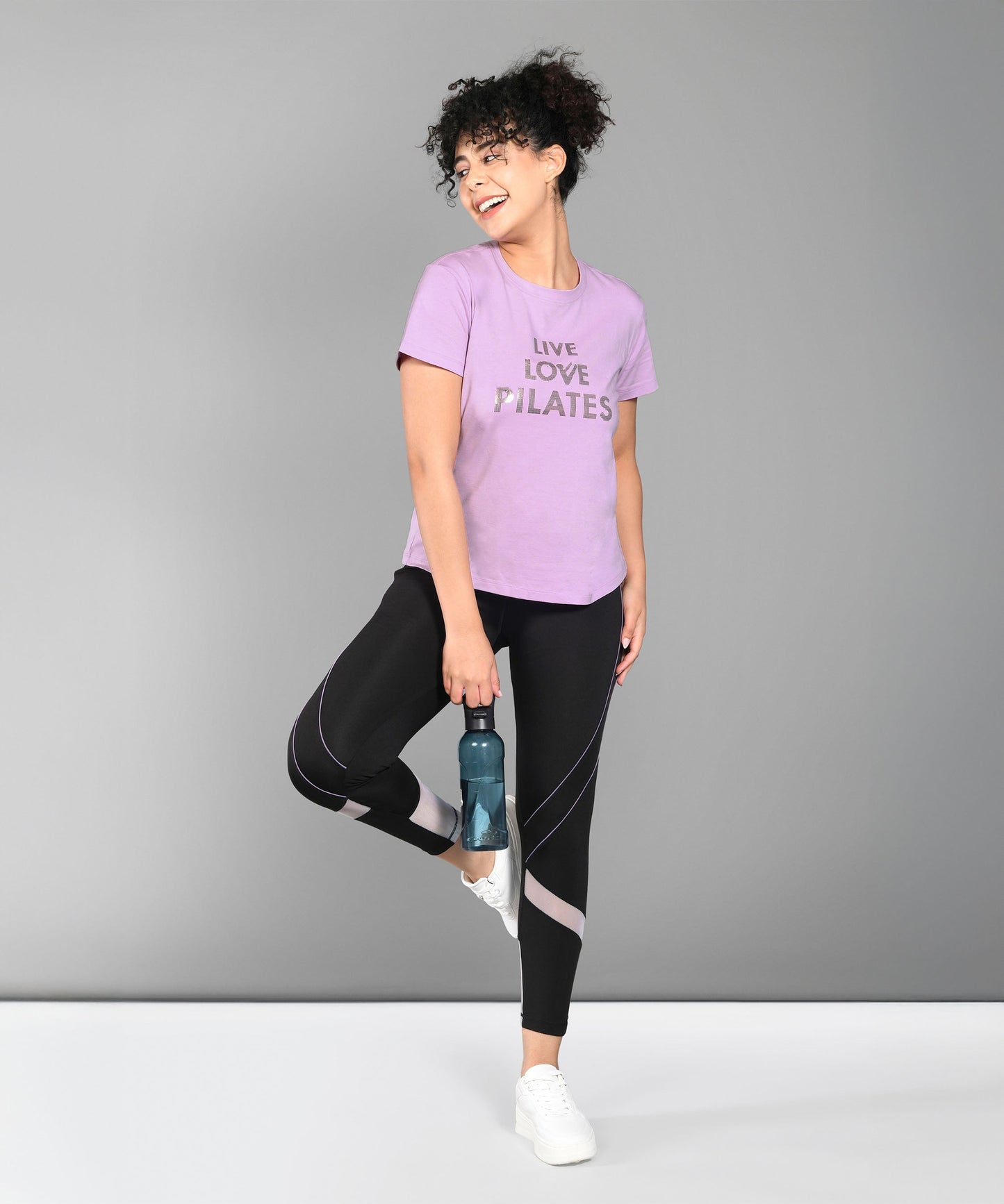 A woman in workout clothes smiles and poses with a water bottle, wearing the Pilates Pink T-shirt—a soft, stylish cotton-modal tee that's perfect for women seeking comfort and flair during Pilates workouts.
