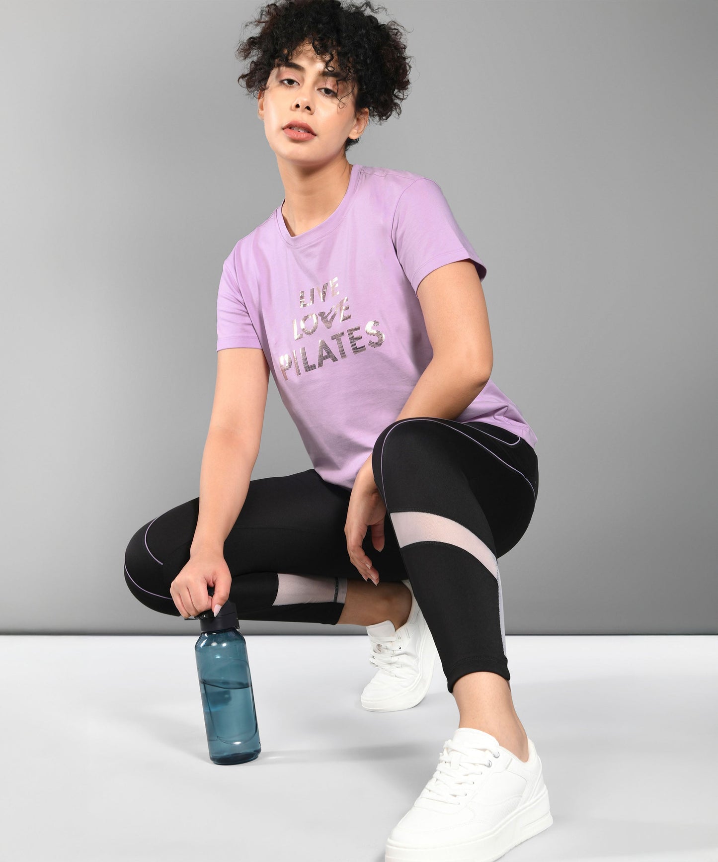 A person squats in activewear, holding a blue water bottle and wearing our Pilates Pink T-shirt from the cotton modal tee collection, featuring "Live Love Pilates" on the front.