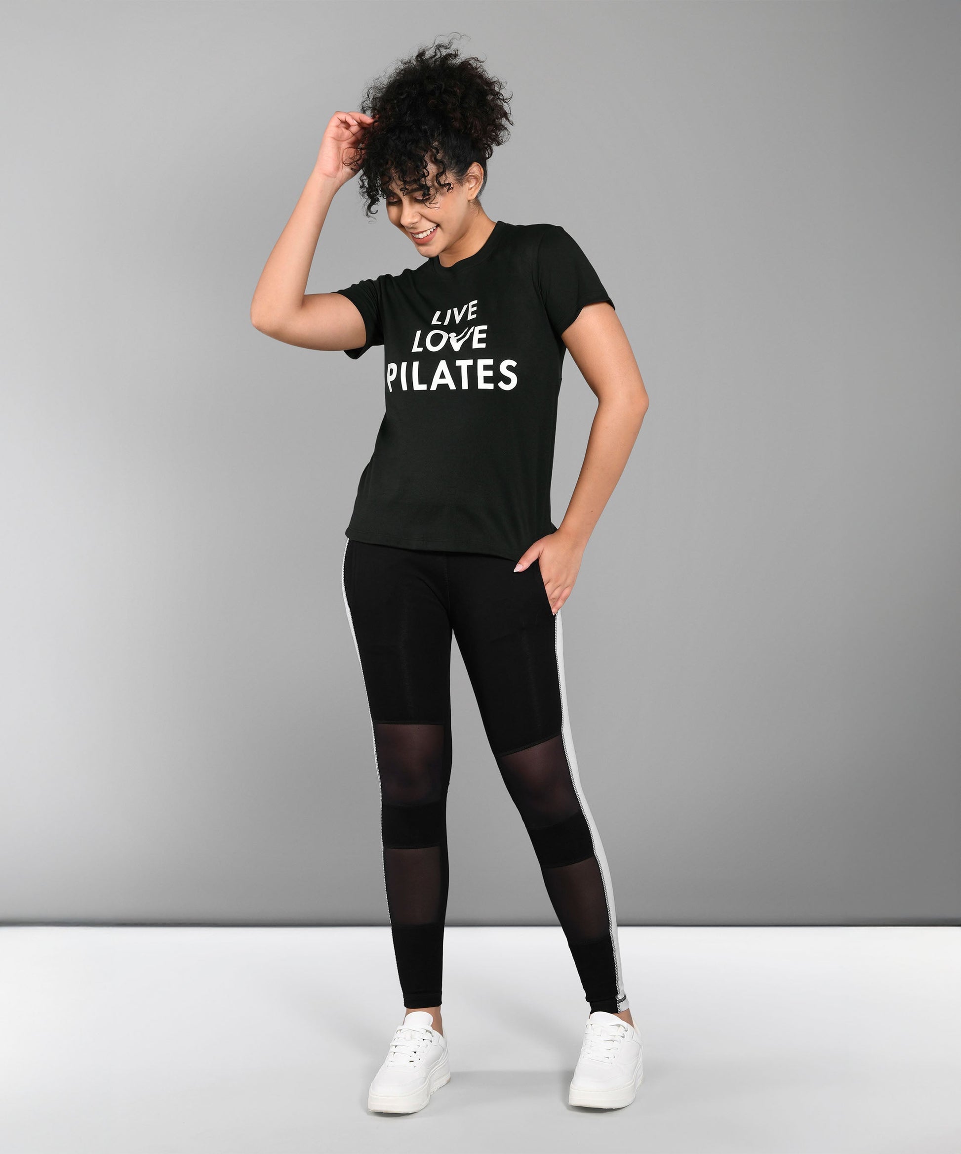 Smiling woman in a Pilates Black T-shirt and leggings stands against a gray background.
