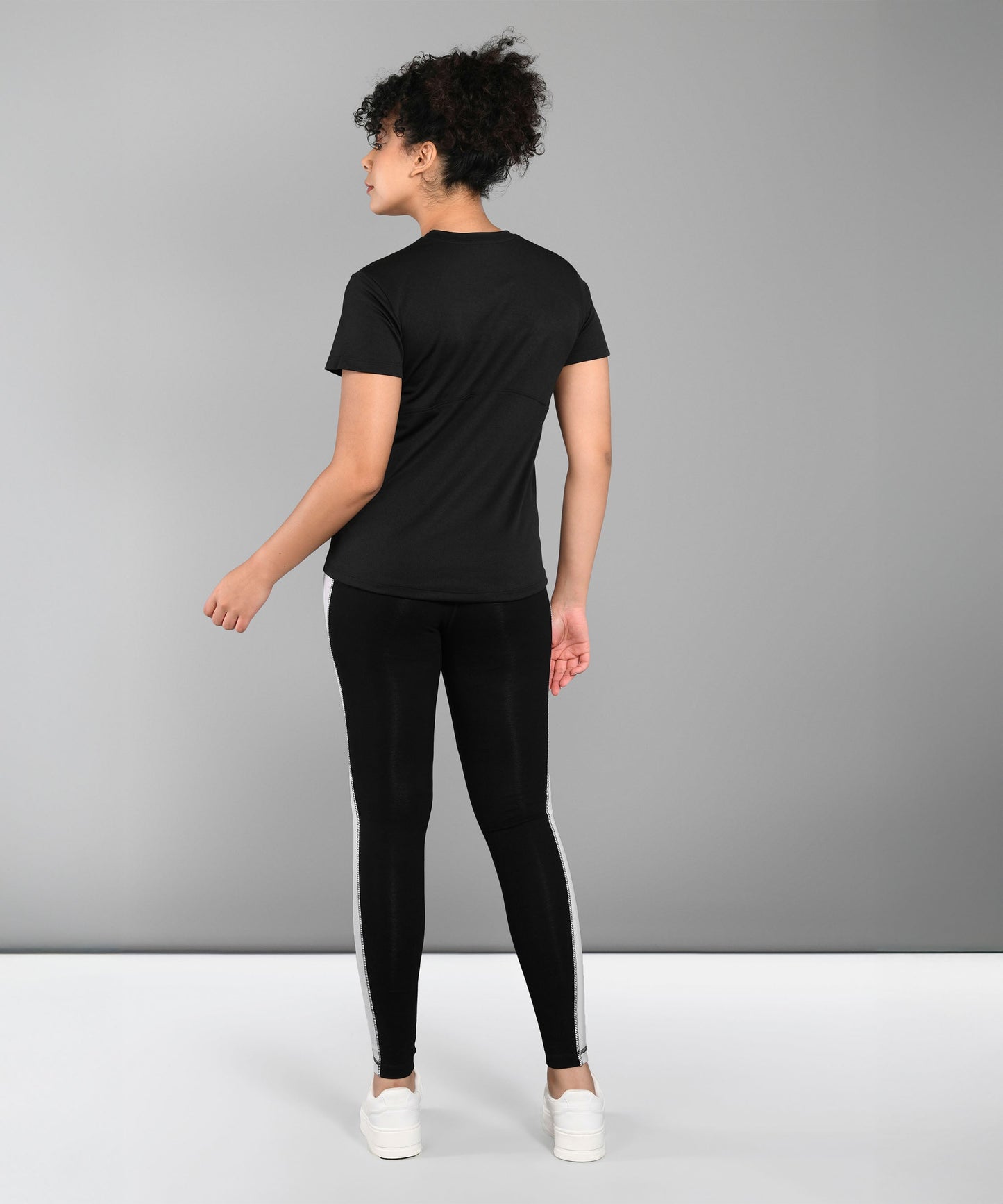 A person stands facing away, wearing a Pilates Black T-shirt, black leggings with white stripes, and white sneakers.