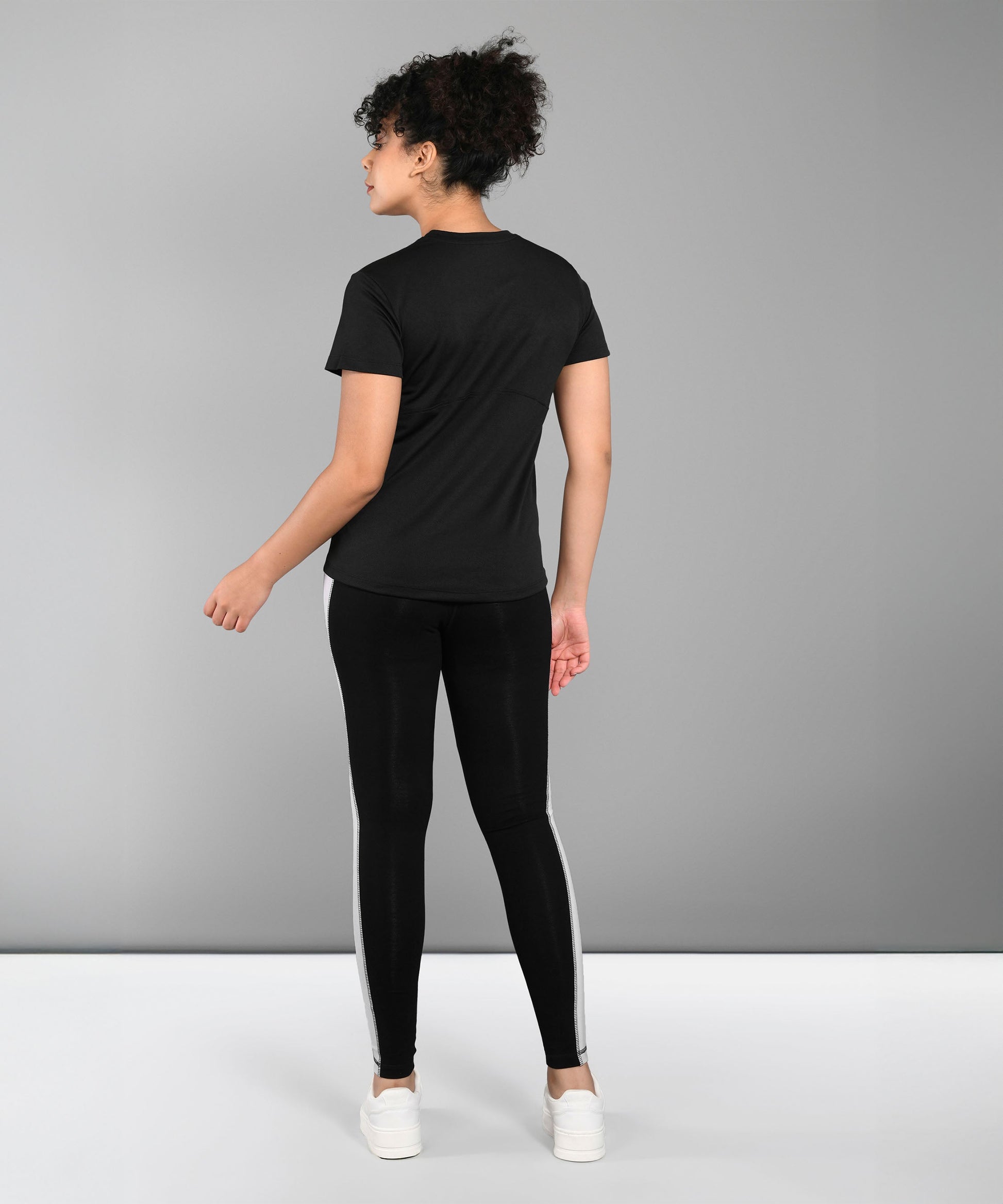 A person stands facing away, wearing a Pilates Black T-shirt, black leggings with white stripes, and white sneakers.