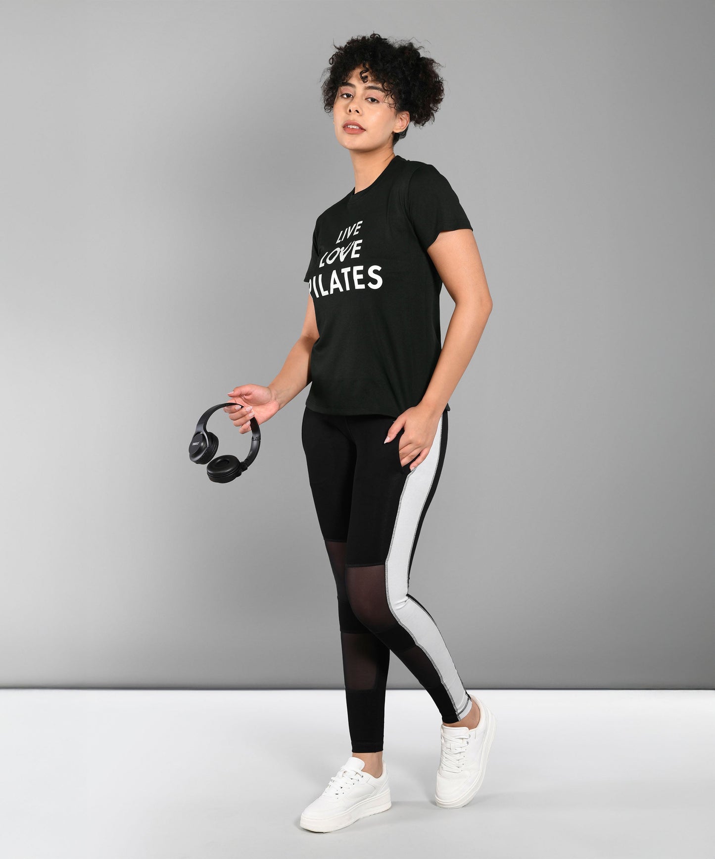 Woman in activewear holding headphones, wearing the Pilates Black T-shirt featuring a "Live Love Pilates" print, standing against a plain background.