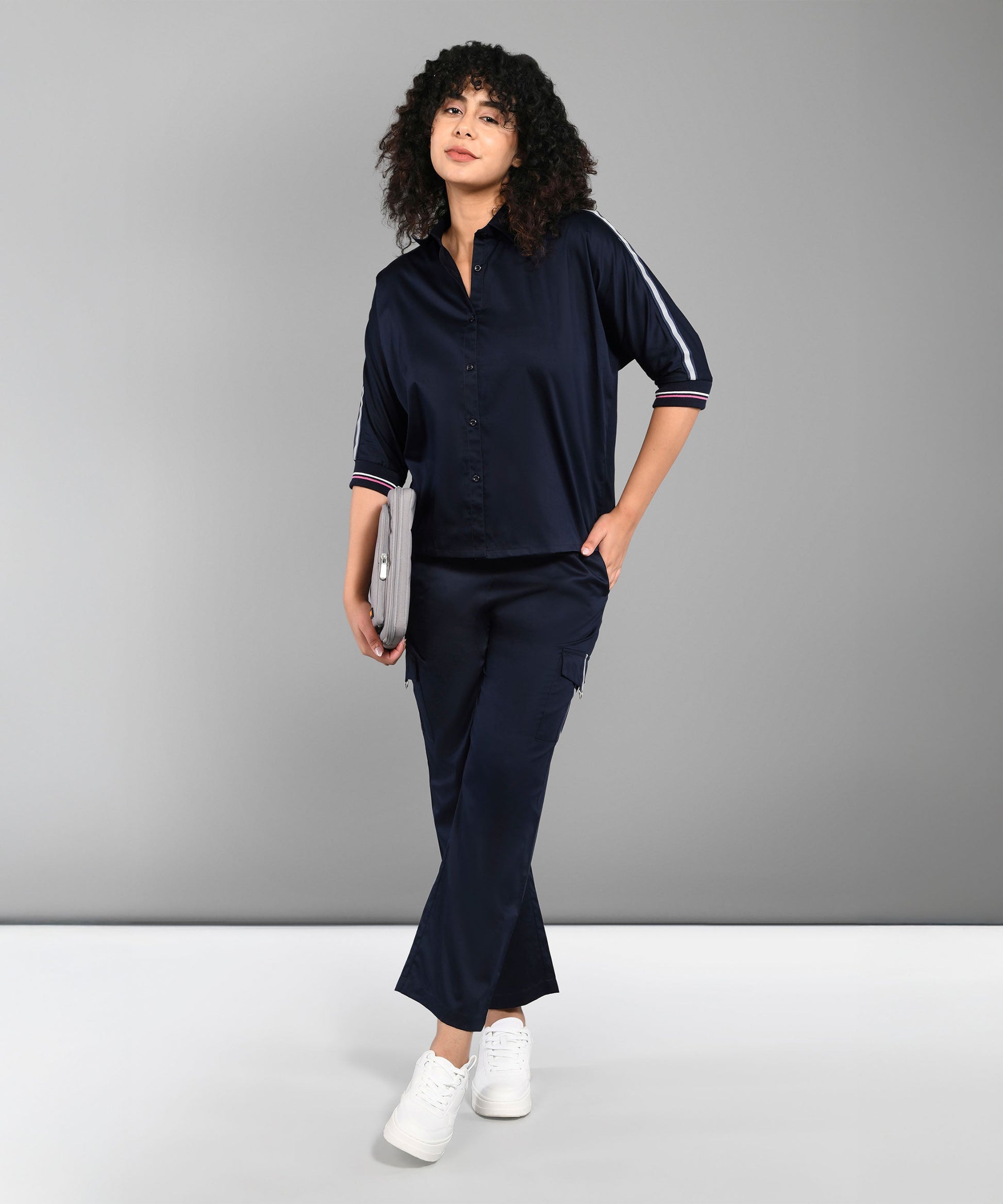 Woman with curly hair in the Navy Blue Chic Set and white sneakers, holding a tablet and standing against a gray background.