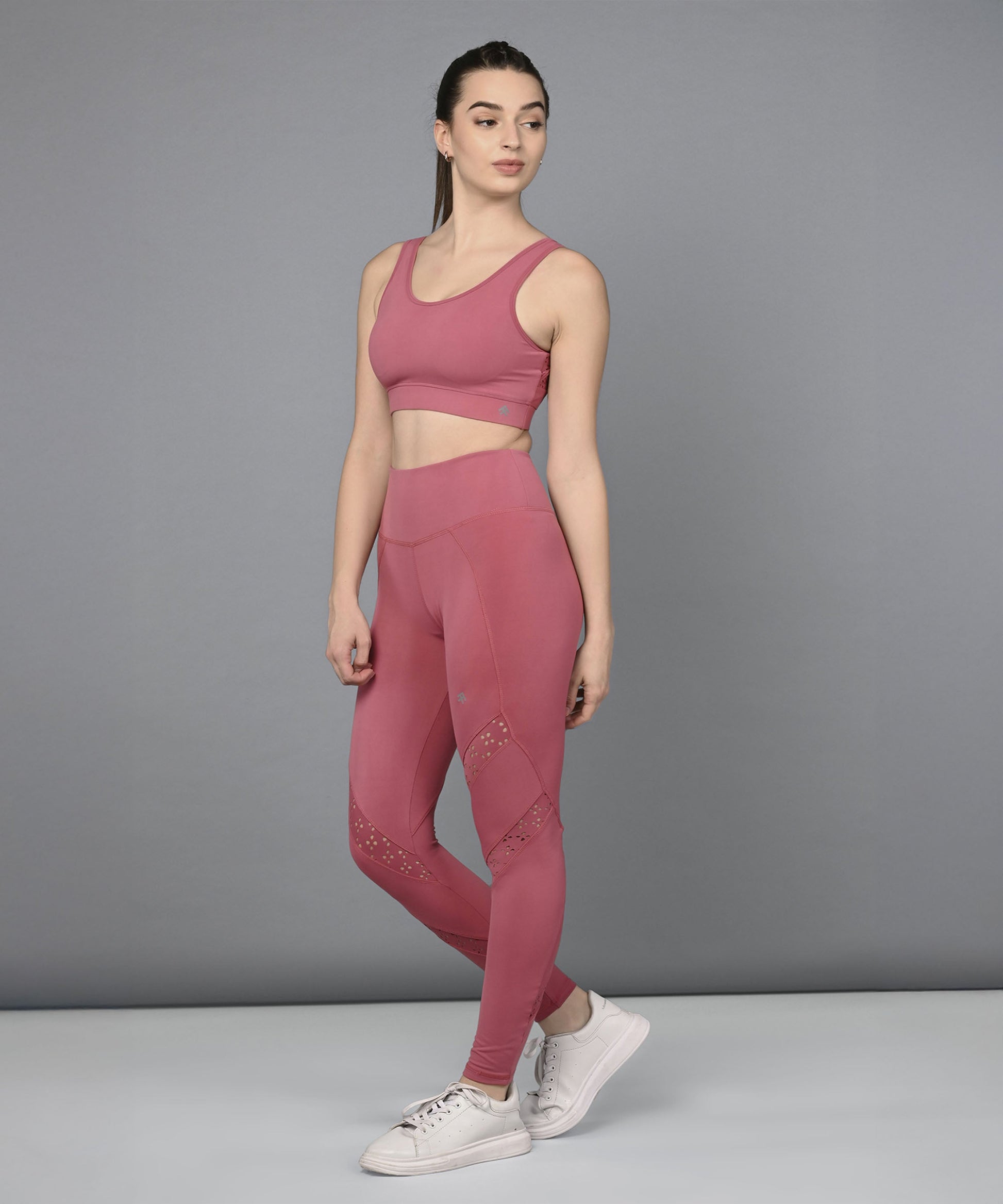 A woman wearing the Peony Pink Netted Top and matching leggings with white sneakers stands against a plain gray background.