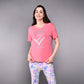 A woman smiles, wearing the 'Adore Your Core' Printed Tee in pink with vibrant patterned leggings against a gray background. This motivational shirt adds a touch of positivity to any fitness routine.