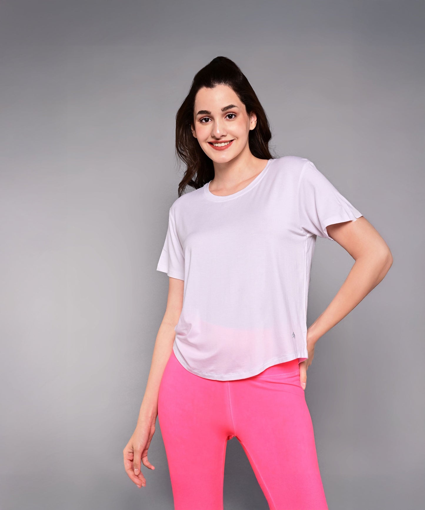 A woman in a light pink 'Pilates Has Your Back' Printed Tee and bright pink leggings smiles confidently against a gray background—the ideal motivational shirt for any Pilates tee collection.
