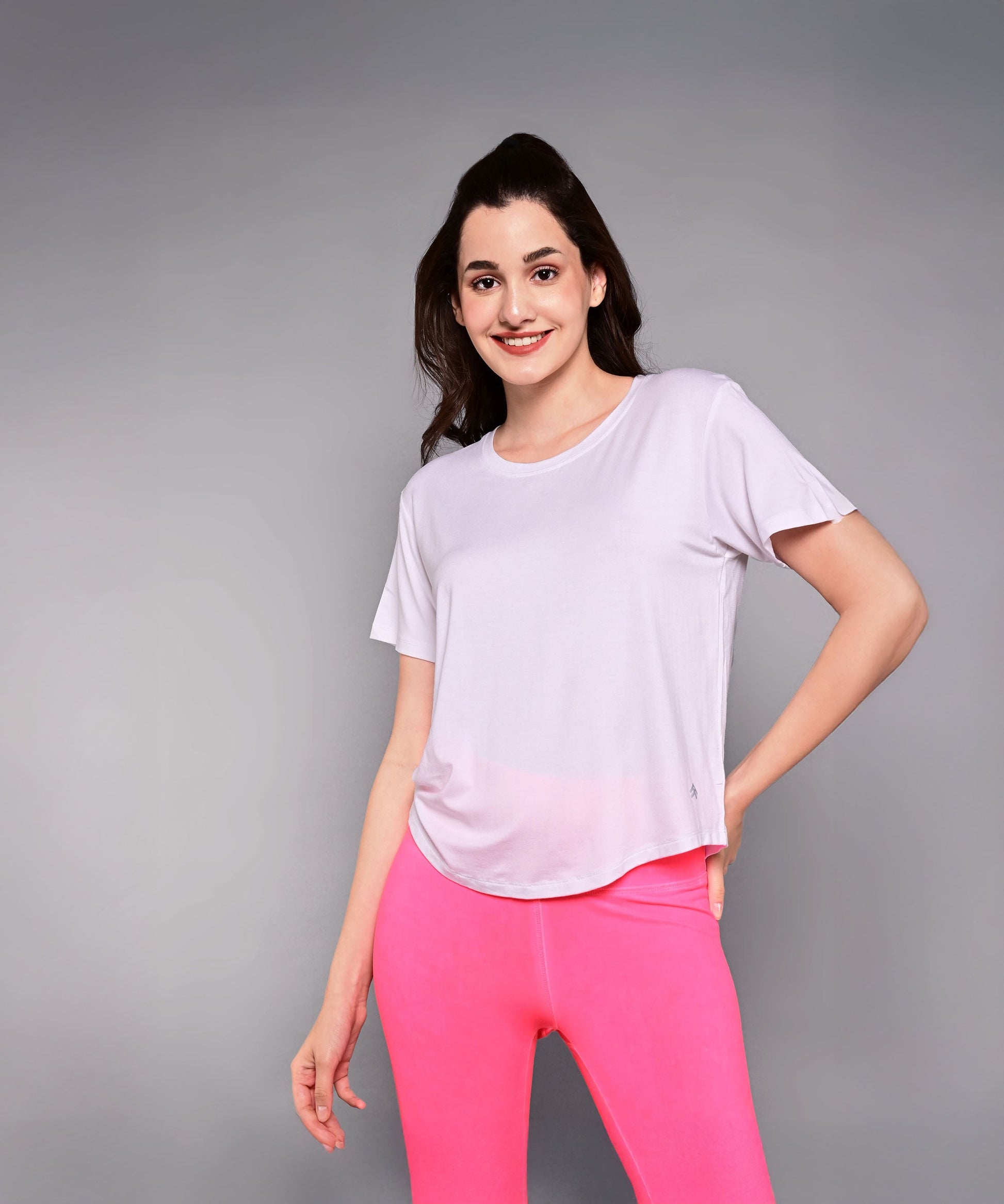 A woman in a light pink 'Pilates Has Your Back' Printed Tee and bright pink leggings smiles confidently against a gray background—the ideal motivational shirt for any Pilates tee collection.