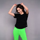 A woman smiles with hands raised, wearing the 'Pilates Has Your Back' Printed Tee and bright green leggings against a gray background.