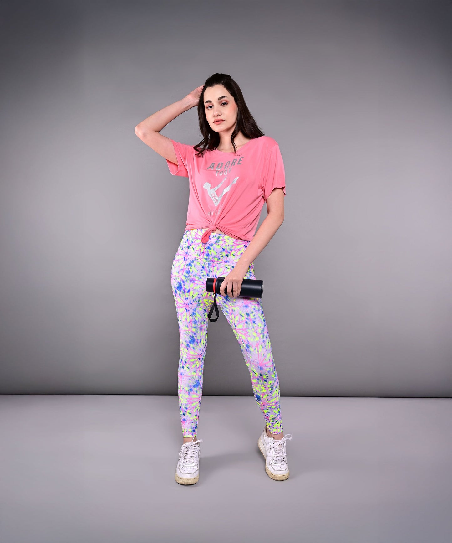 A woman poses against a gray background, holding a camera and wearing the 'Adore Your Core' Printed Tee with colorful leggings—her look radiates energy and confidence.