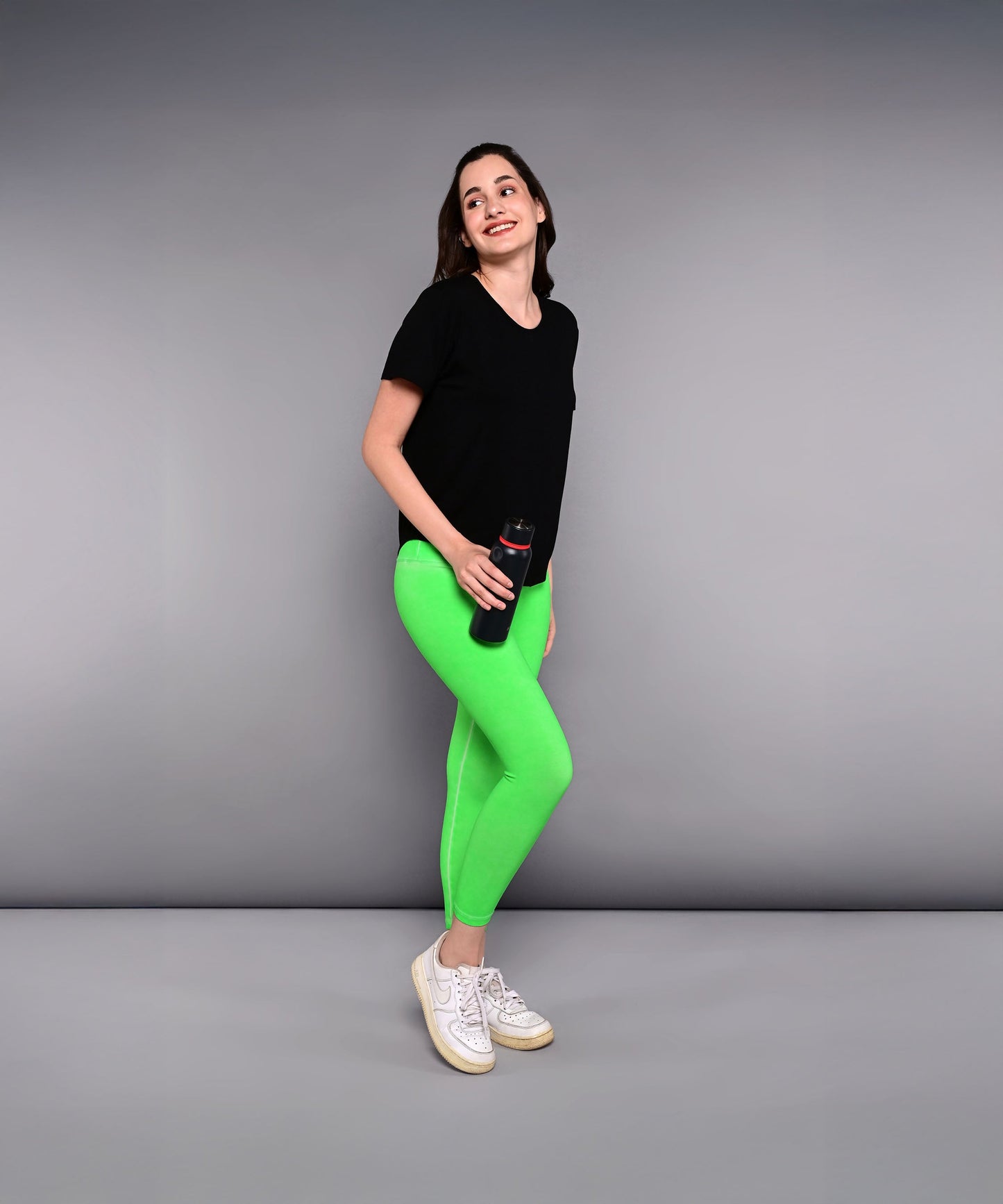 Woman in a 'Pilates Has Your Back' printed tee and bright green leggings holding a water bottle, standing against a gray background.