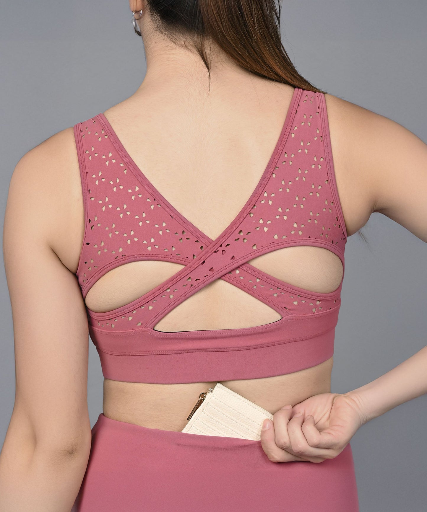 A woman wearing the Peony Pink Sports Bra with a cutout back holds a small wallet behind her. This moisture-wicking fitness top keeps you cool and comfortable during workouts.
