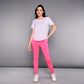 A woman stands against a gray background wearing the 'Pilates Has Your Back' Printed Tee—a white cotton modal shirt—paired with bright pink leggings and white sneakers.