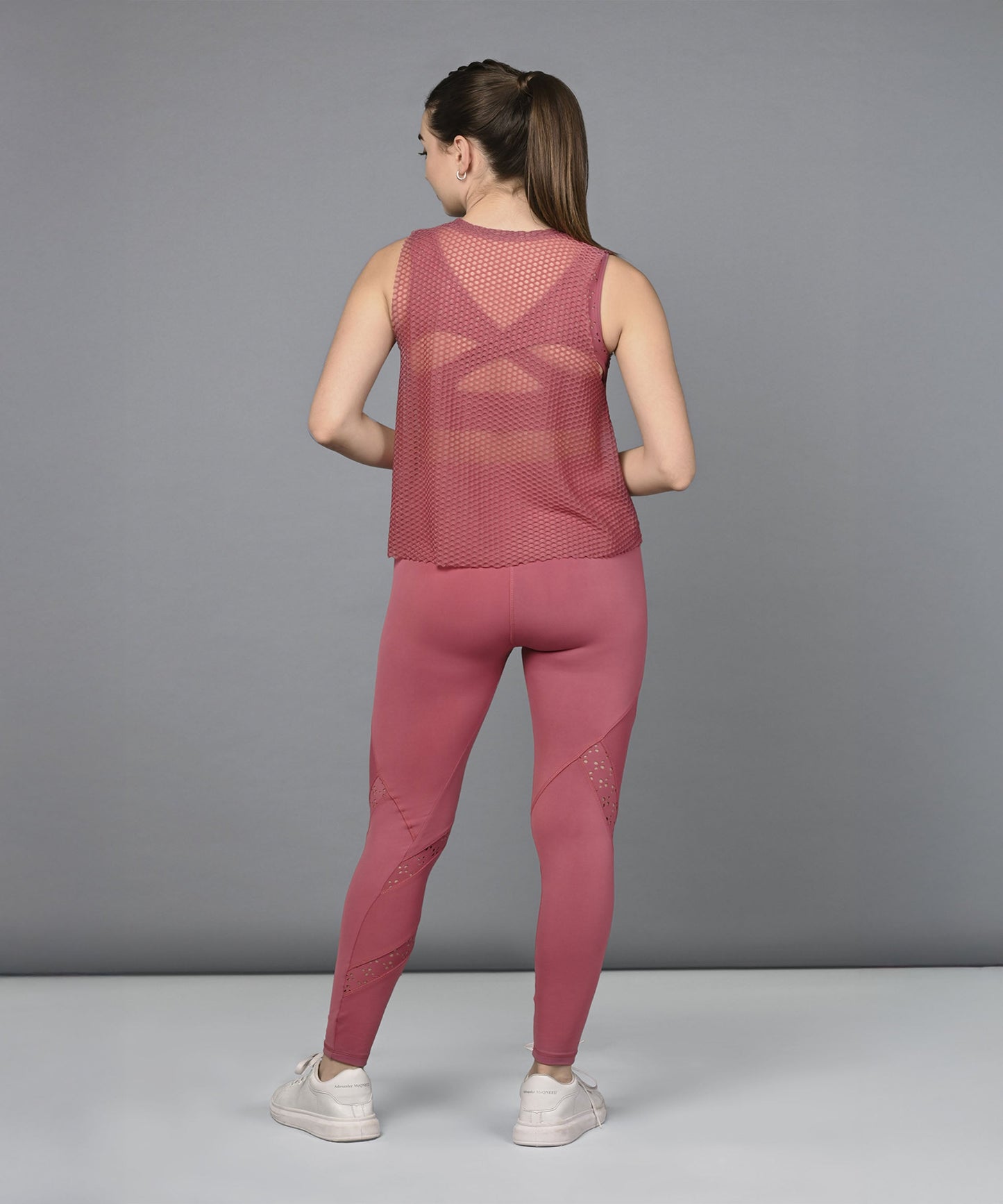 A woman stands facing away in the Peony Pink Netted Top, displaying its mesh design and honeycomb self pattern against a gray background.