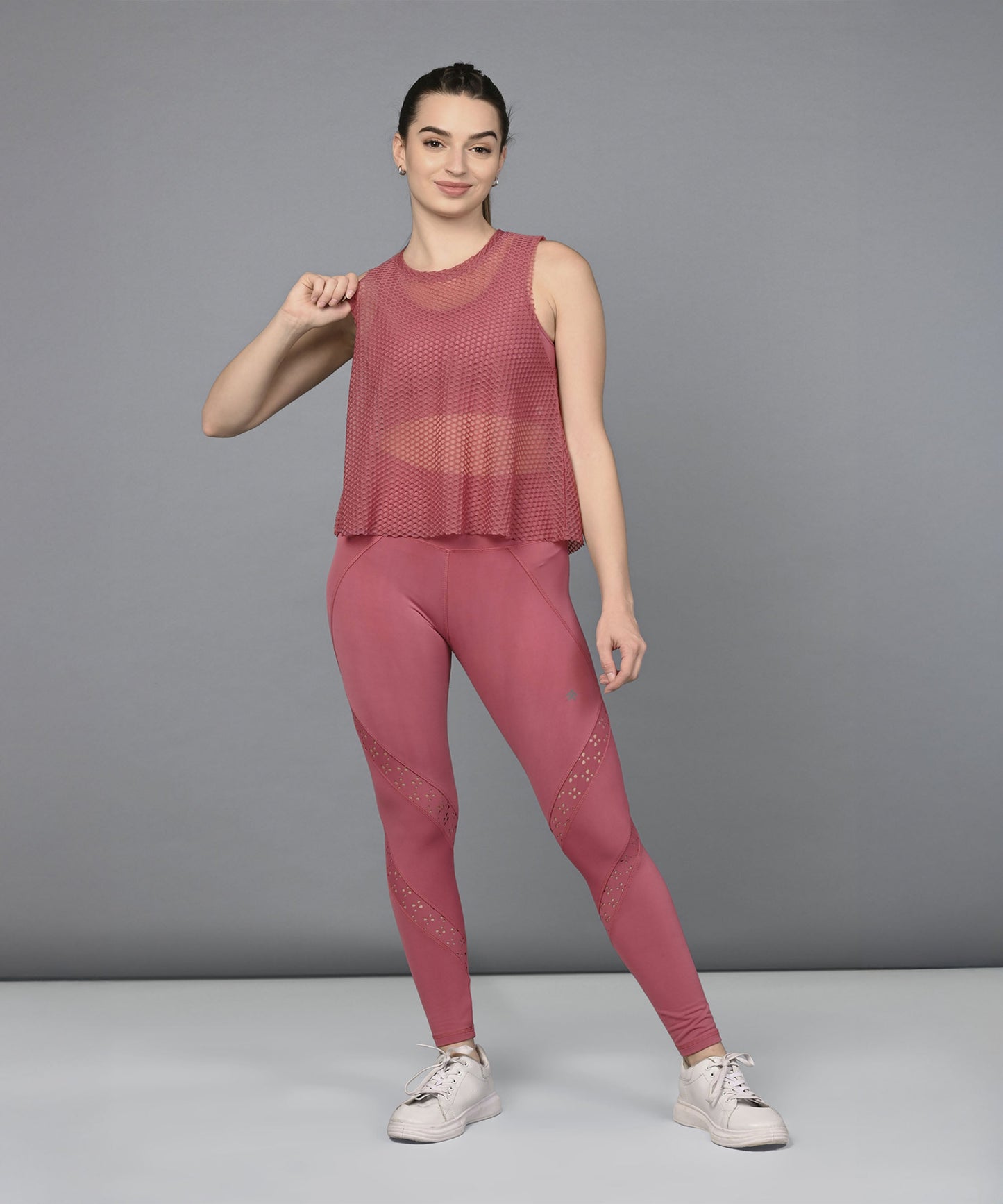 A woman poses confidently against a gray background, showcasing the Peony Pink Netted Top paired with white sneakers for a chic, sporty look.