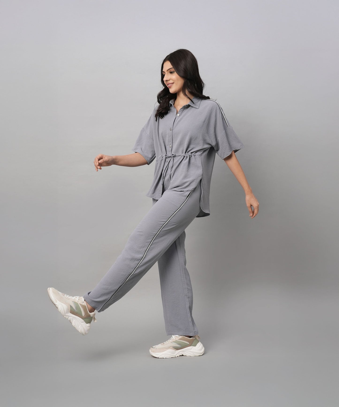 A woman wears the Grey Tie Up Top with Loose Pants Cord Set, pairing the coordinated grey outfit with white sneakers as she walks against a plain grey background.