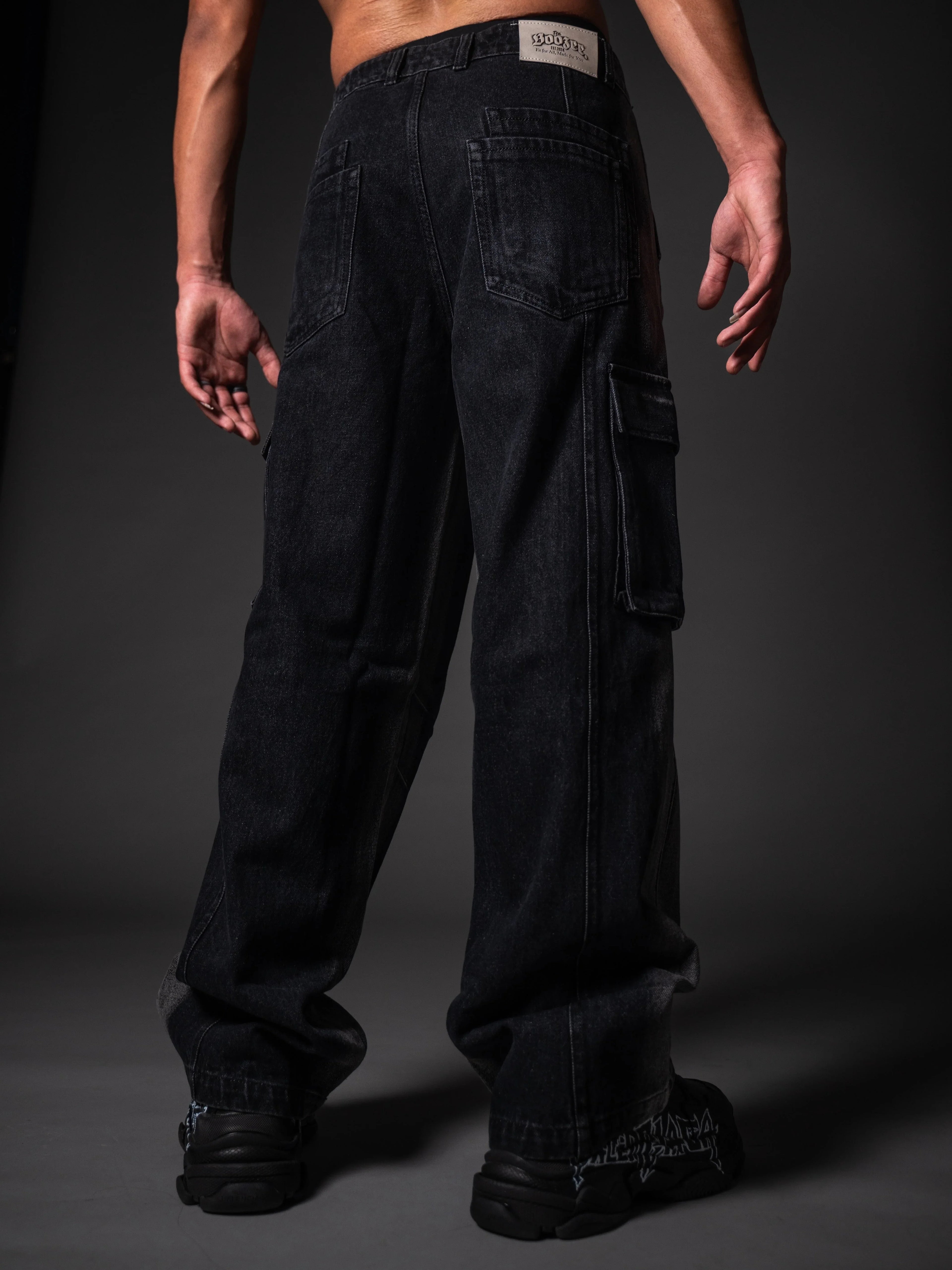 A back view of a model wearing loose-fitting, black denim cargo pants. The jeans feature standard back pockets, a side cargo pocket on the right leg, and a white branded patch on the right waistband. The look is completed with chunky black sneakers featuring white graphic details, set against a dark grey studio background.