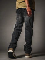 A back view of a model wearing relaxed-fit, faded black denim jeans. The jeans feature a vintage wash and distinctive back pockets with angled top stitching and flaps. The look is completed with an olive green shirt and low-top sneakers in black, cream, and brown, set against a dark grey studio background