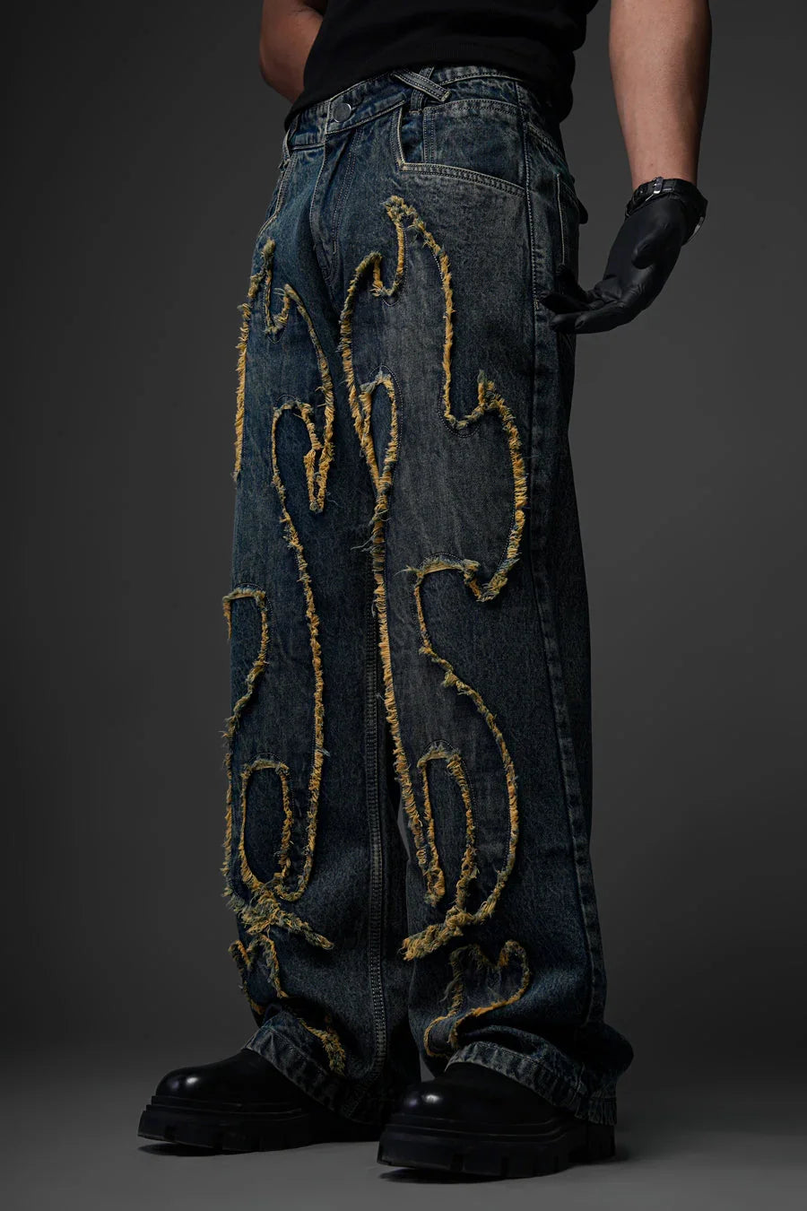 A person from the waist down modeling a pair of baggy, dark blue denim jeans. The jeans feature a bold, raised, and frayed pattern shaped like flames running down both legs, with yellowish-tan frayed edges. They are paired with chunky black leather boots and a black leather glove.