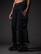 A model from the waist down wearing loose-fitting, faded black denim cargo pants with large side pocket flaps. The outfit is paired with chunky black sneakers featuring white graffiti-style lettering on the sides, set against a dark grey studio background.
