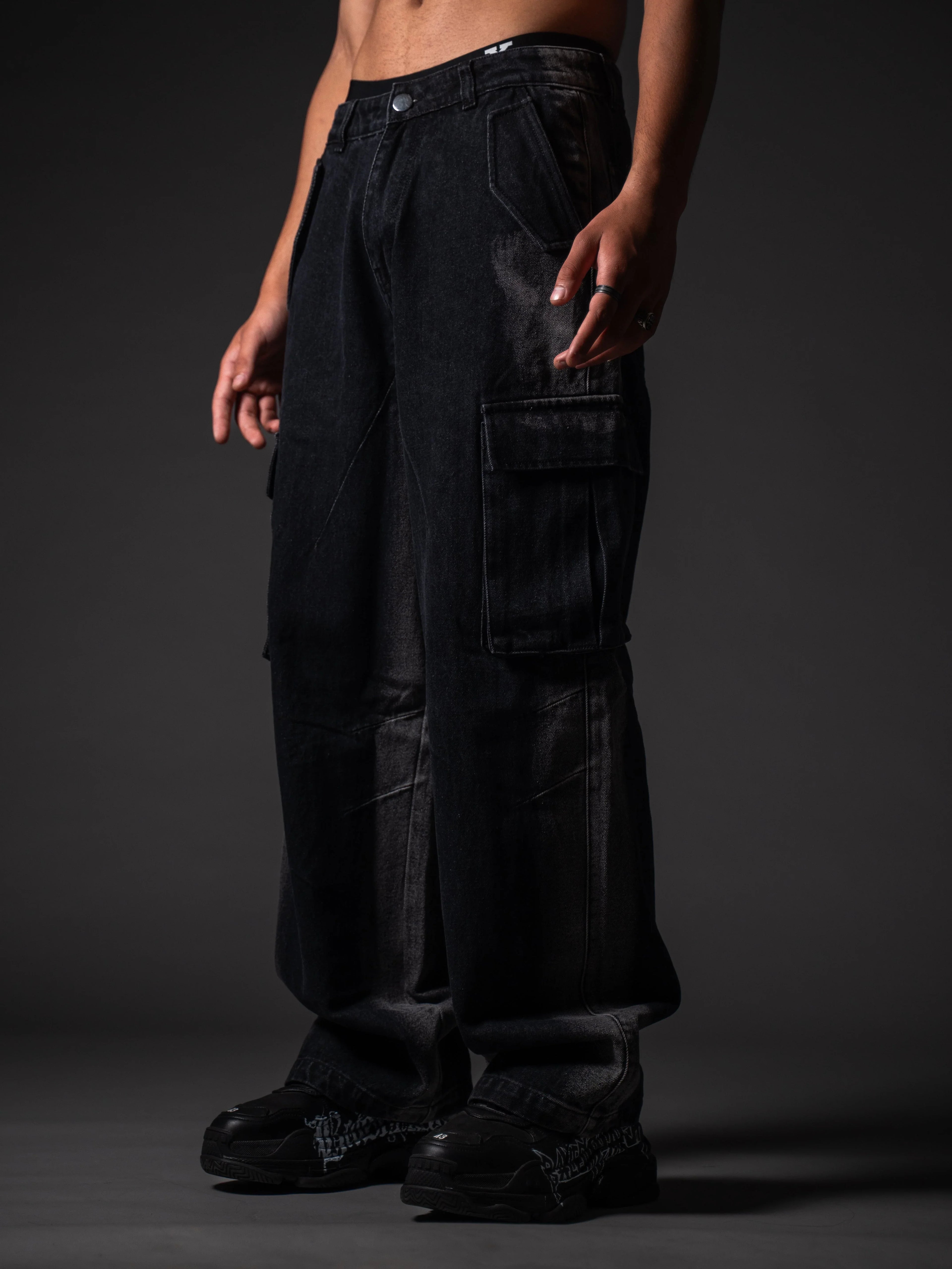 A model from the waist down wearing loose-fitting, faded black denim cargo pants with large side pocket flaps. The outfit is paired with chunky black sneakers featuring white graffiti-style lettering on the sides, set against a dark grey studio background.