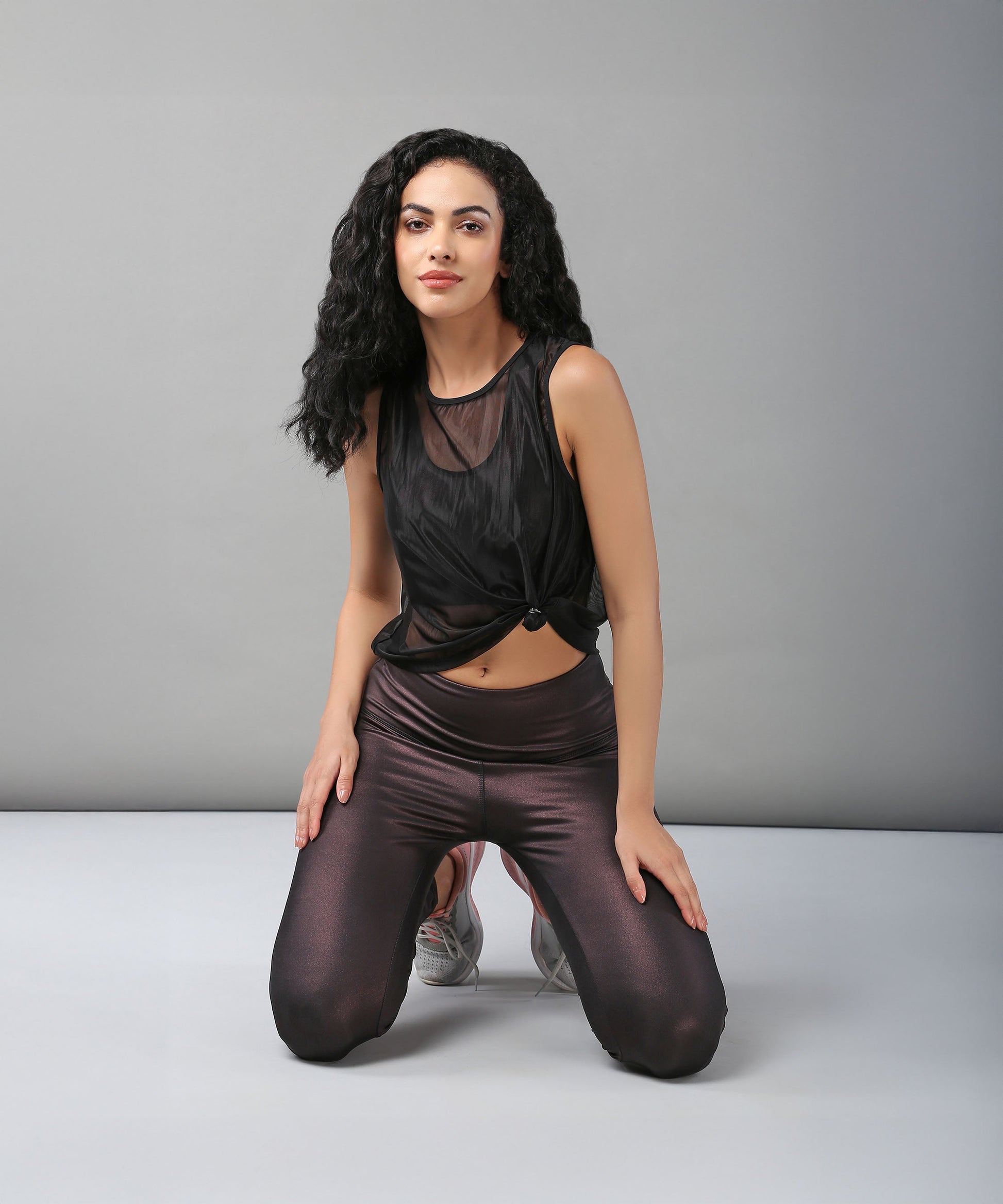 Woman with long curly hair kneeling on the floor, wearing a sexy Black Sheer Top and shiny dark leggings.