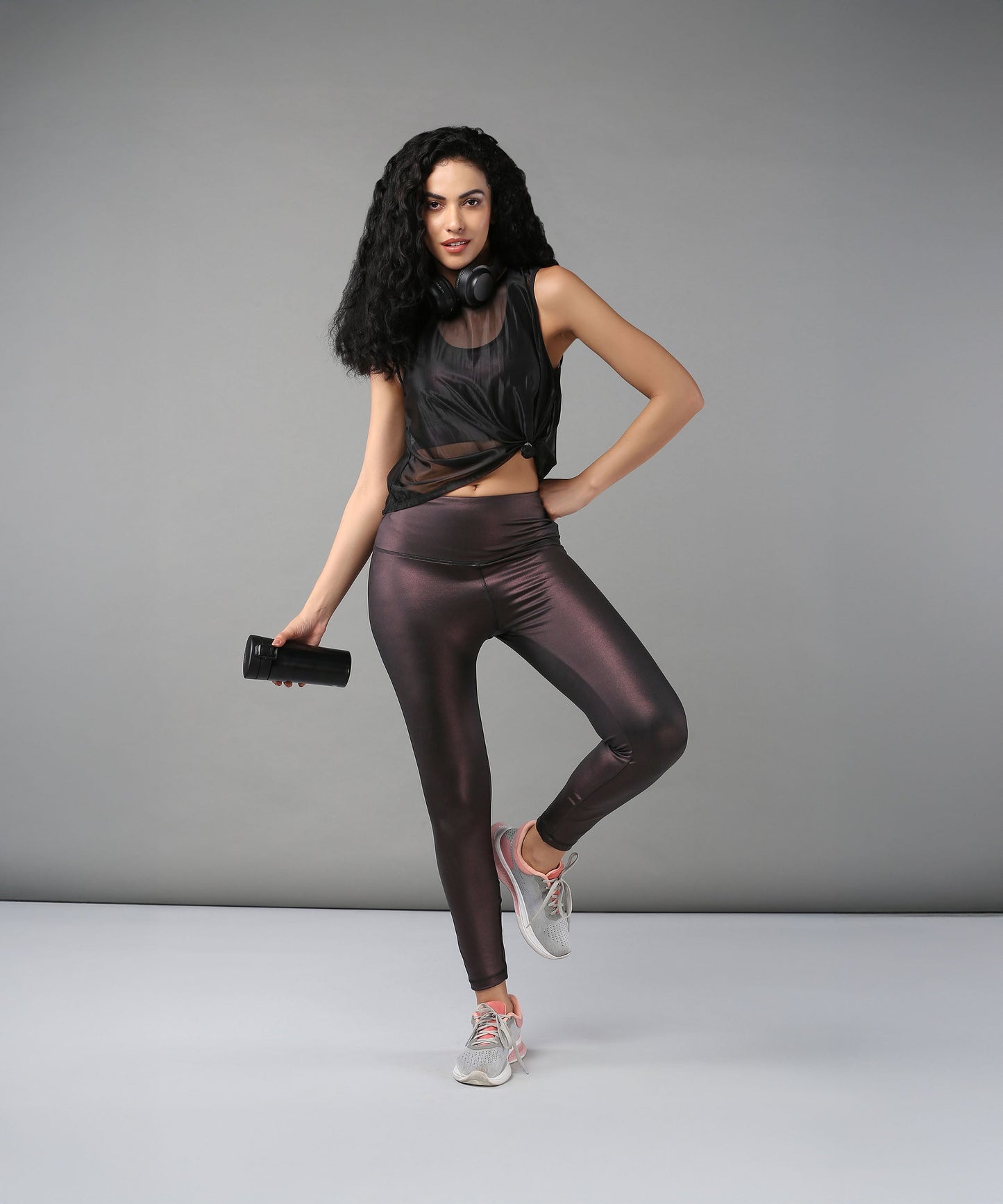 Confident woman poses against a gray background wearing headphones, a Black Sheer Top, shiny workout clothes, and holding a water bottle.