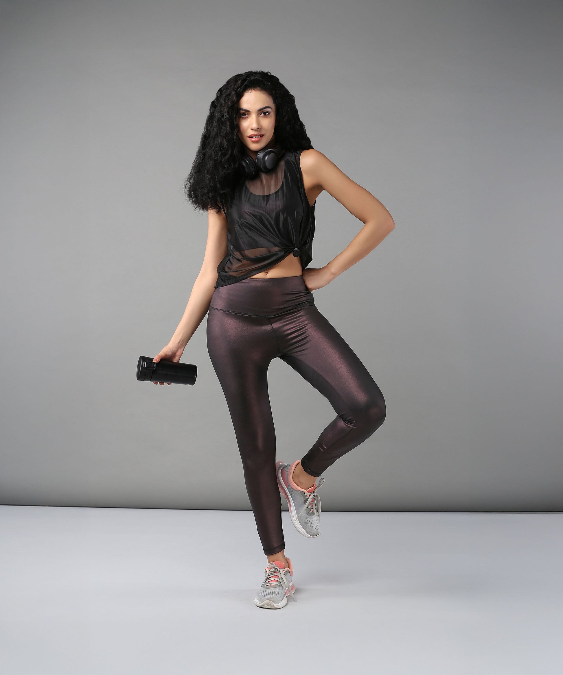 Confident woman poses against a gray background wearing headphones, a Black Sheer Top, shiny workout clothes, and holding a water bottle.