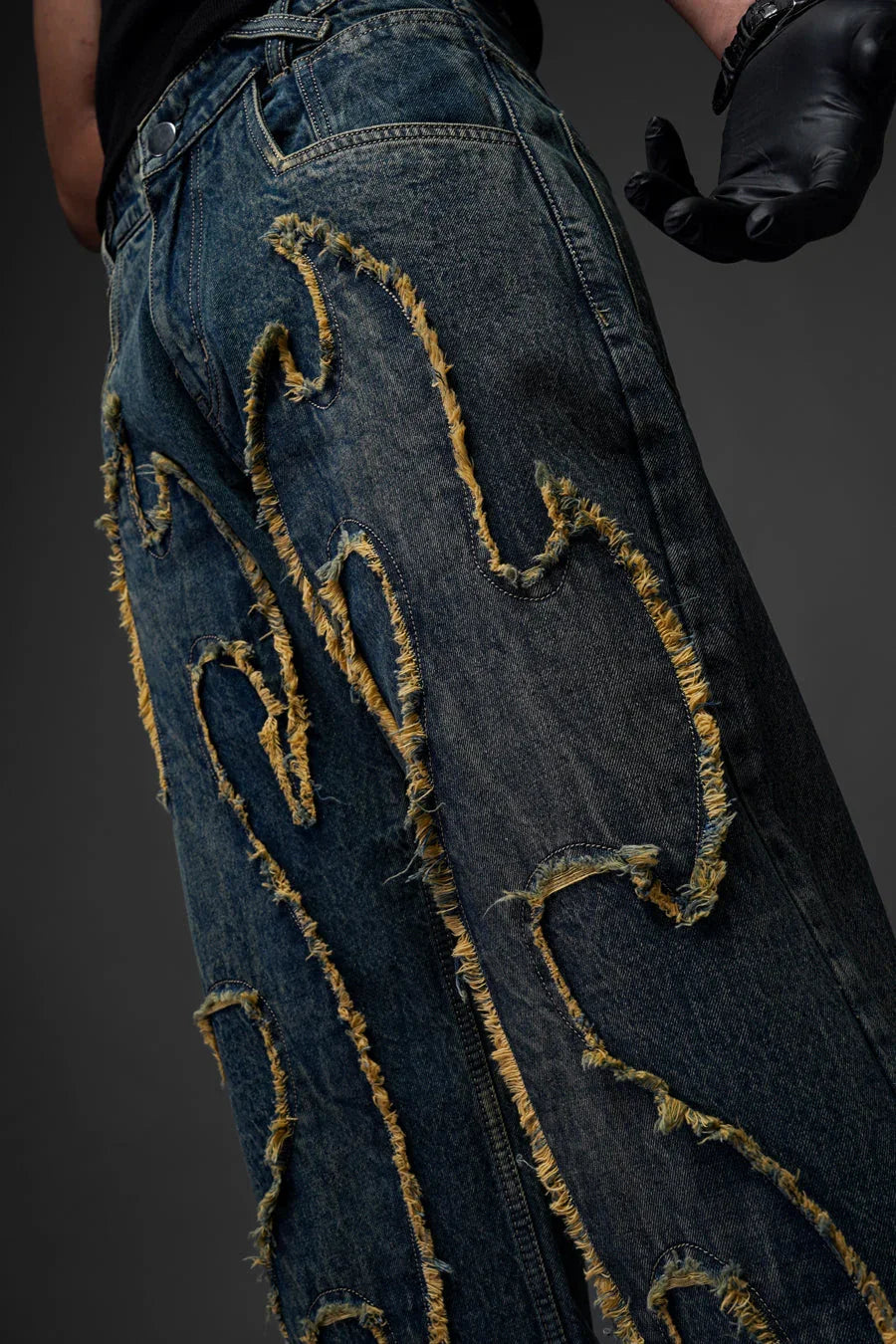 A close-up side view of a person wearing dark blue denim jeans. The focus is on the intricate, raised appliqué design that mimics flames, featuring heavily frayed, yellowish-tan edges. A hand wearing a black leather glove is visible on the right side of the frame.