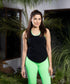 Woman in a Solid Black Top with Racer Back and neon green leggings stands outdoors amid lush plants, smiling at the camera.